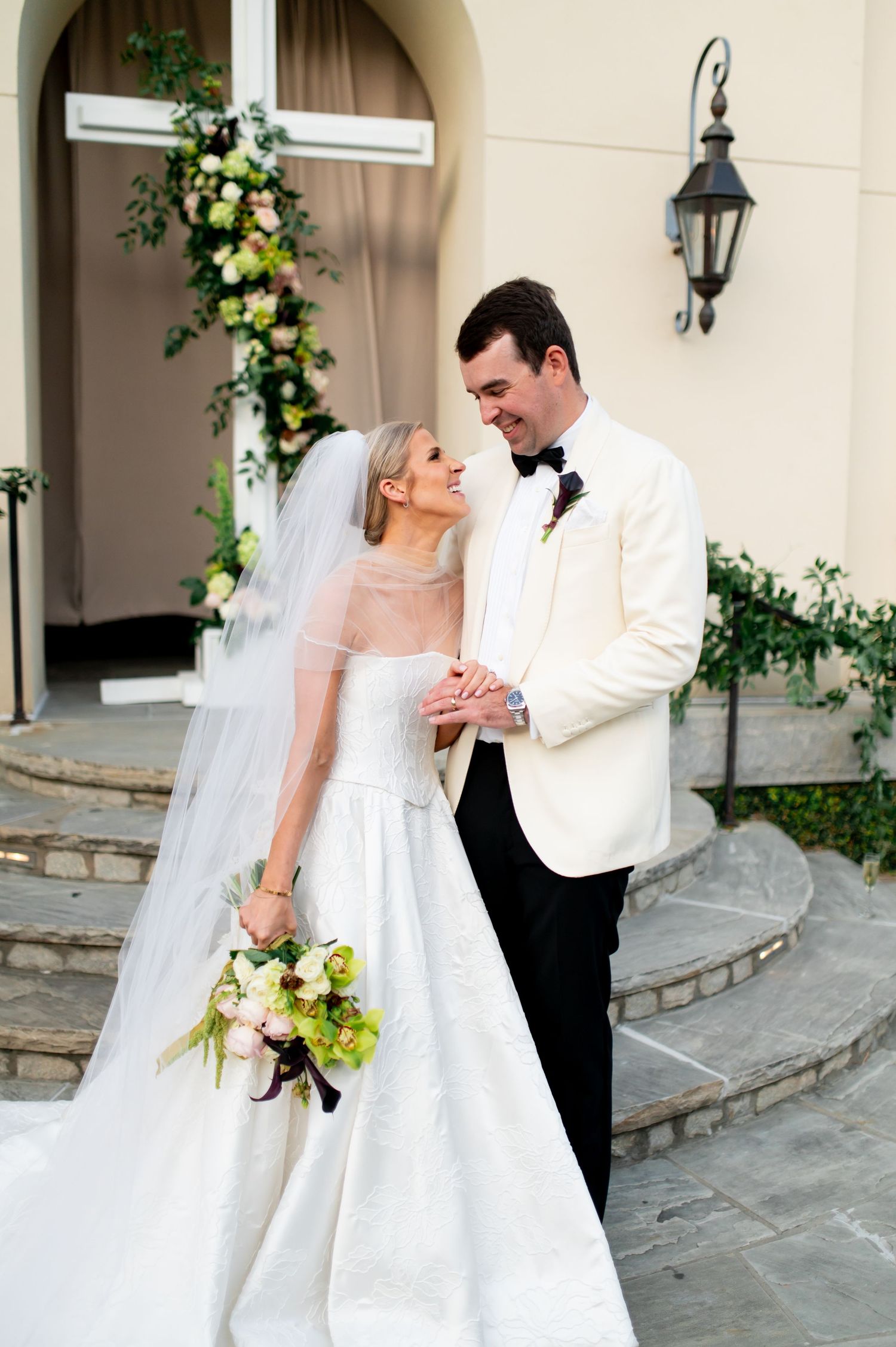 Elegant wedding couple poses against cream building with floral arrangements and lanterns.