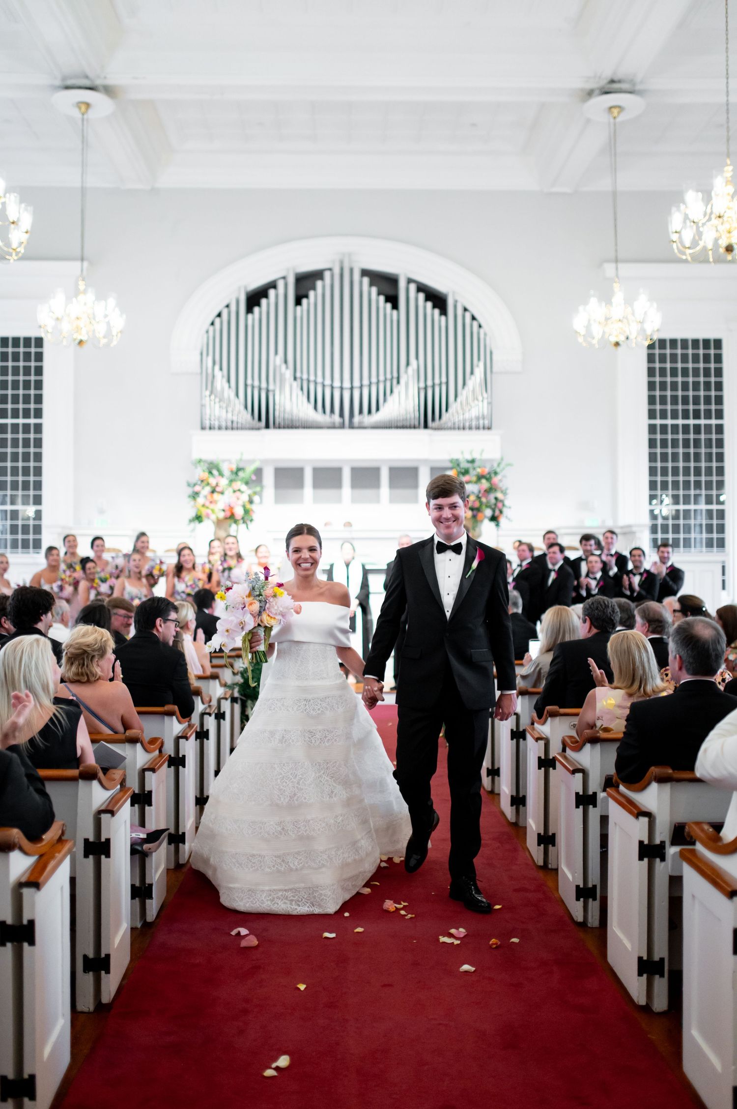 Bride and groom walk down red carpet aisle after ceremony in elegant white church with pipe organ backdrop.