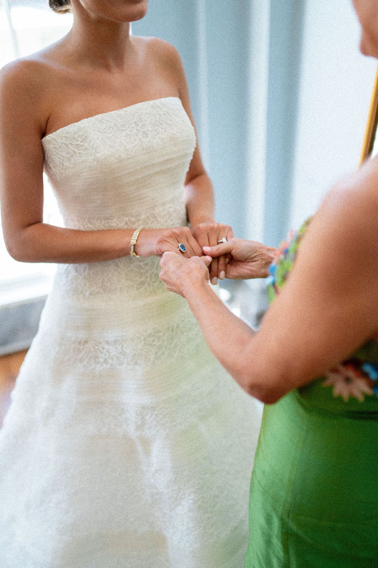 Bridesmaid in green dress fastens bracelet on bride wearing strapless lace wedding gown before ceremony.