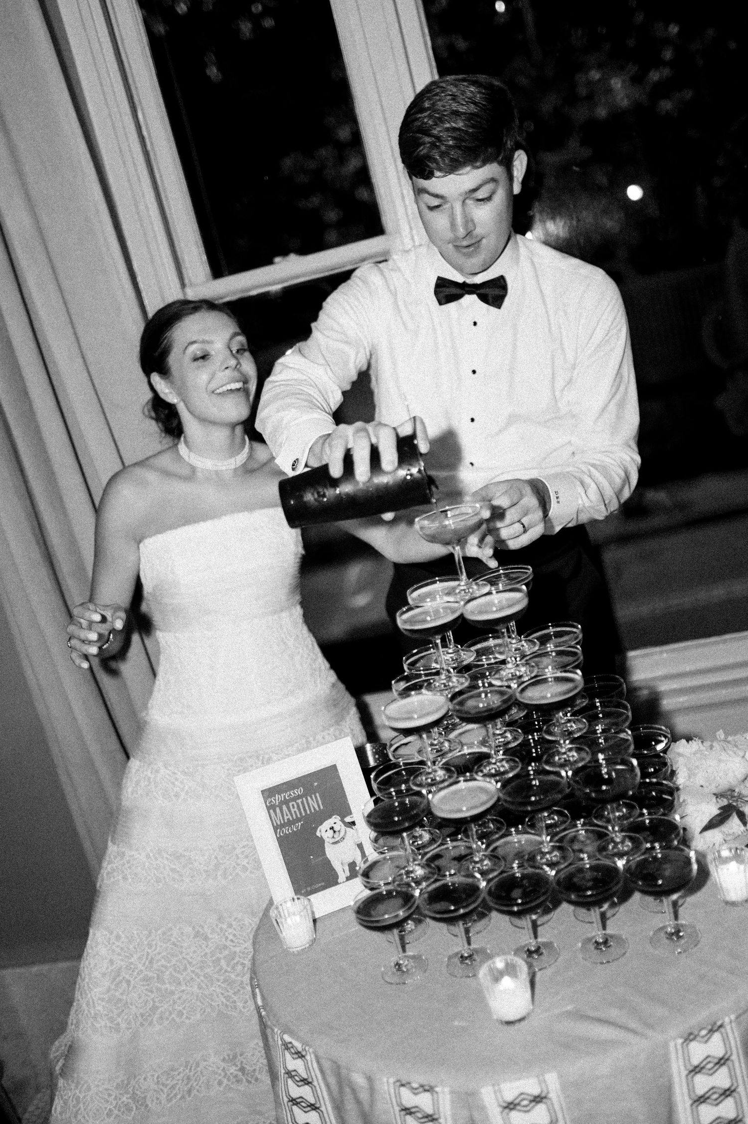 Groom pours champagne tower at wedding reception while bride in strapless gown smiles beside him.