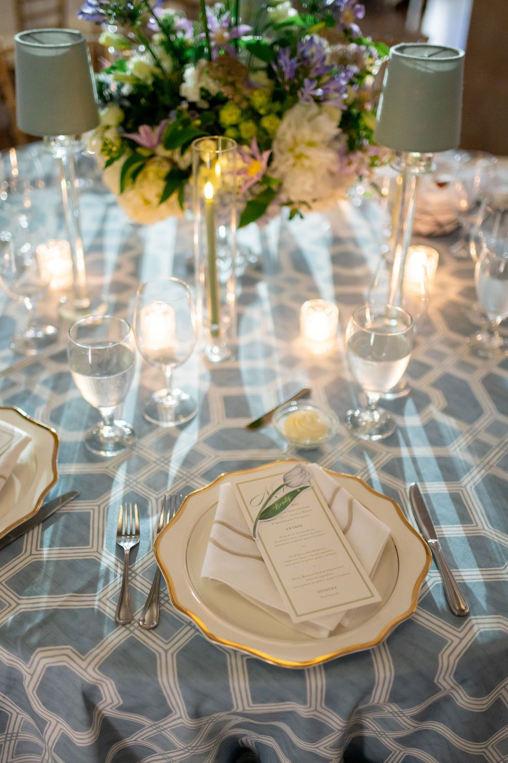 Elegant wedding table setting with gold-rimmed plates, menu cards, candles, and floral centerpiece on blue geometric tablecloth.
