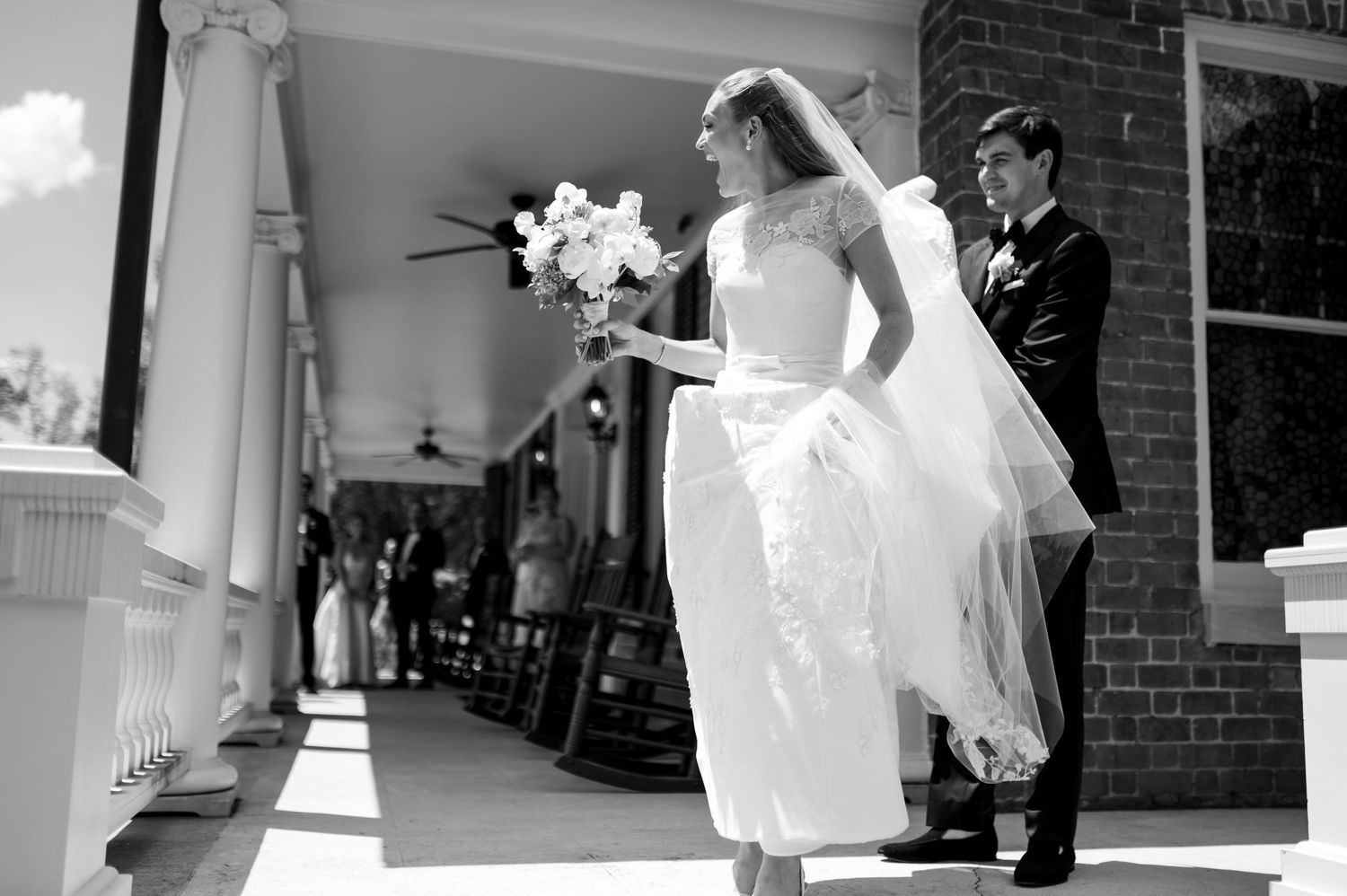 Bride in flowing veil and gown walks with groom on sunlit colonial venue porch in black and white.