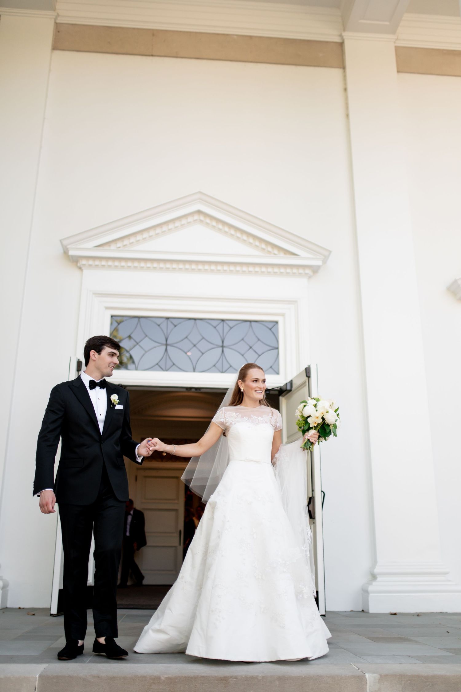 Bride and groom holding hands on church steps, bride in lace cap sleeve gown with veil holding white bouquet.