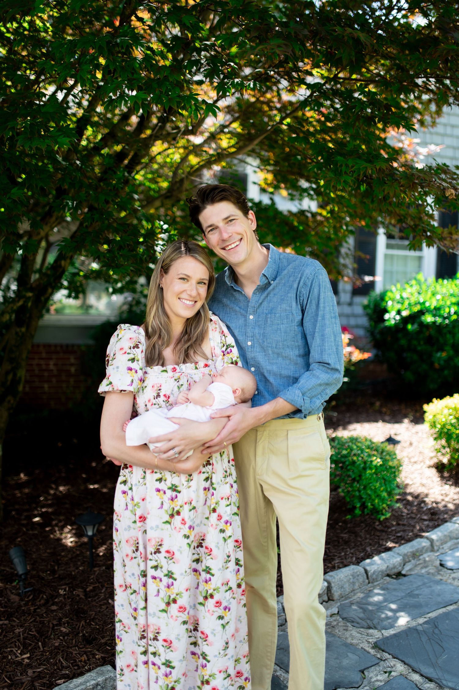 A couple poses with their newborn baby outdoors under a leafy green tree.