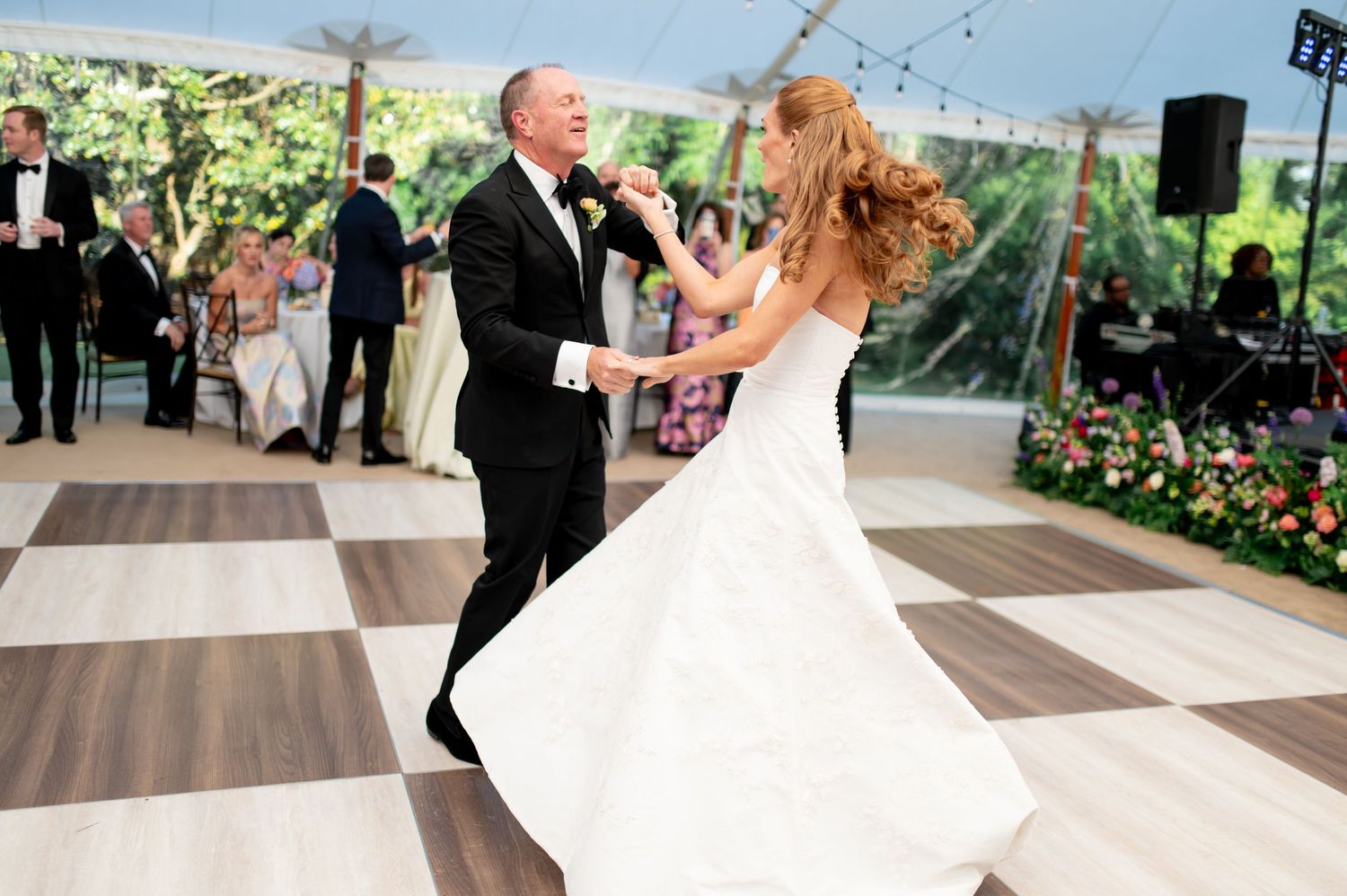 Father and bride share elegant first dance on checkered floor under floral decorated wedding tent.
