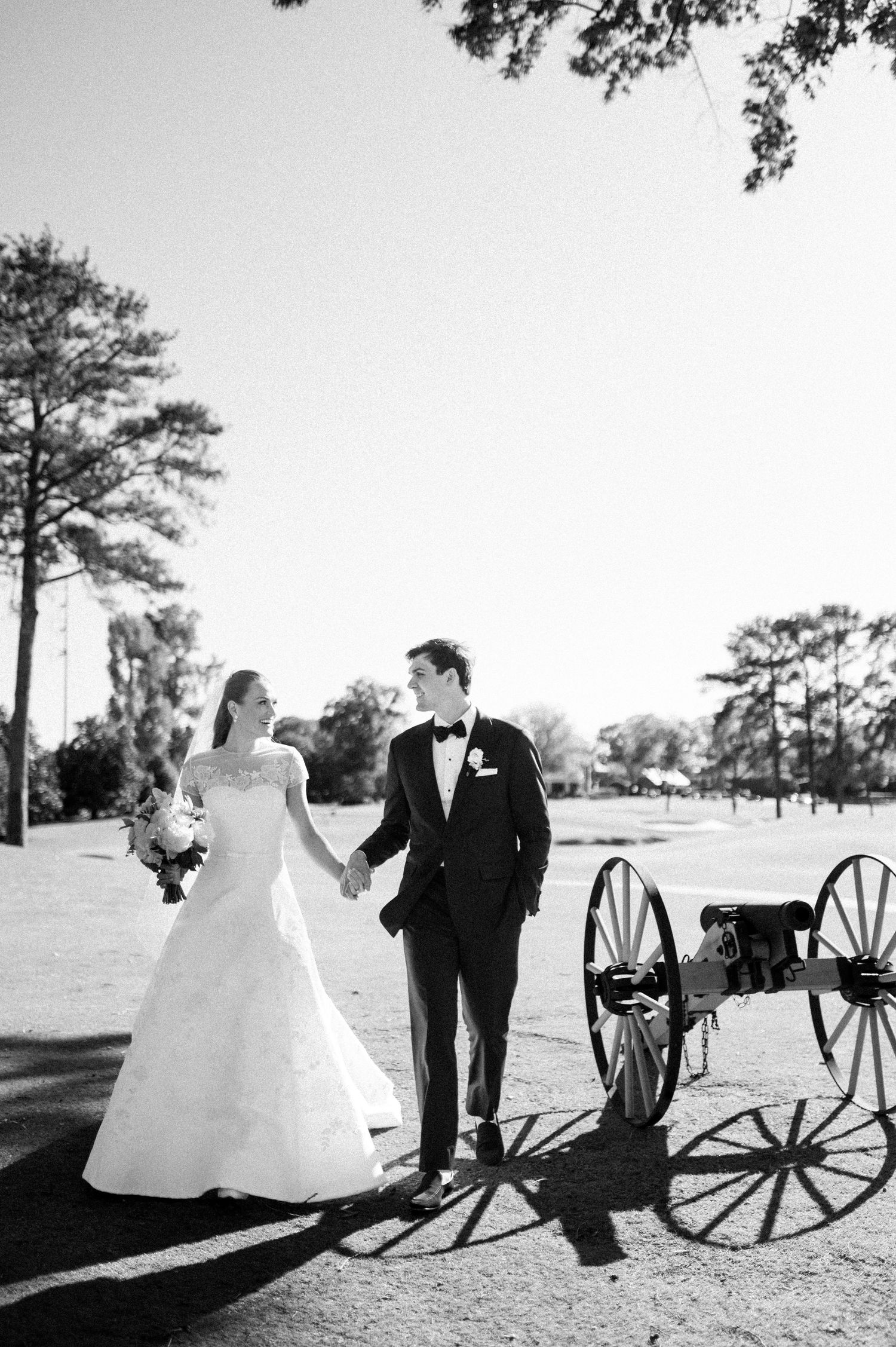 Black and white photo of bride and groom holding hands near vintage cannon on sunny wedding day.