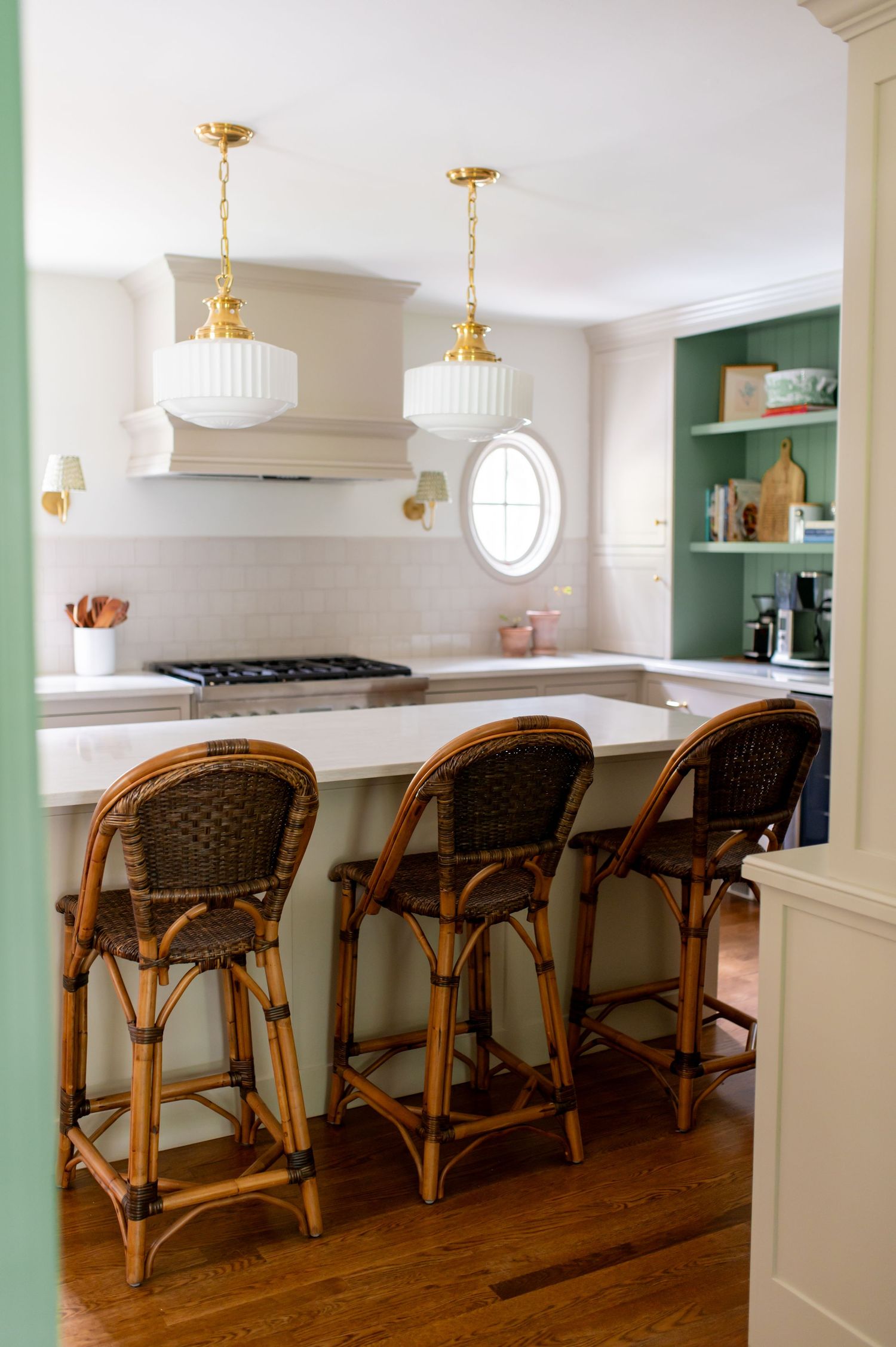 Elegant kitchen interior with rattan bar stools, white countertops, and gold pendant lighting against sage green walls.