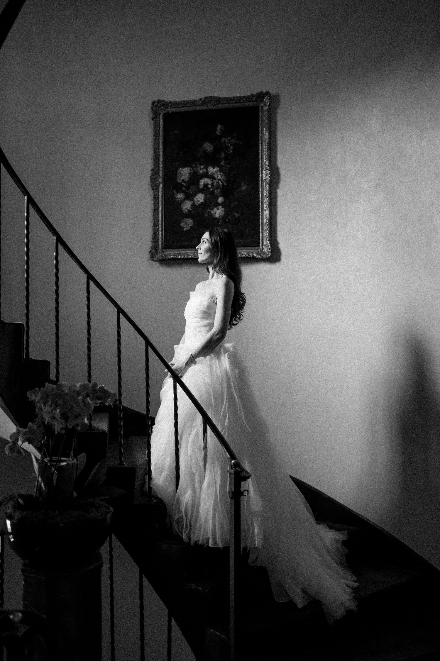 Black and white photo of a bride in a flowing white gown standing on a staircase beneath a framed floral painting.