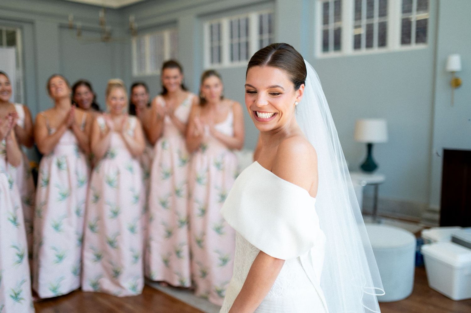Radiant bride in white off-shoulder dress and veil smiles as bridesmaids in floral gowns cheer behind her.