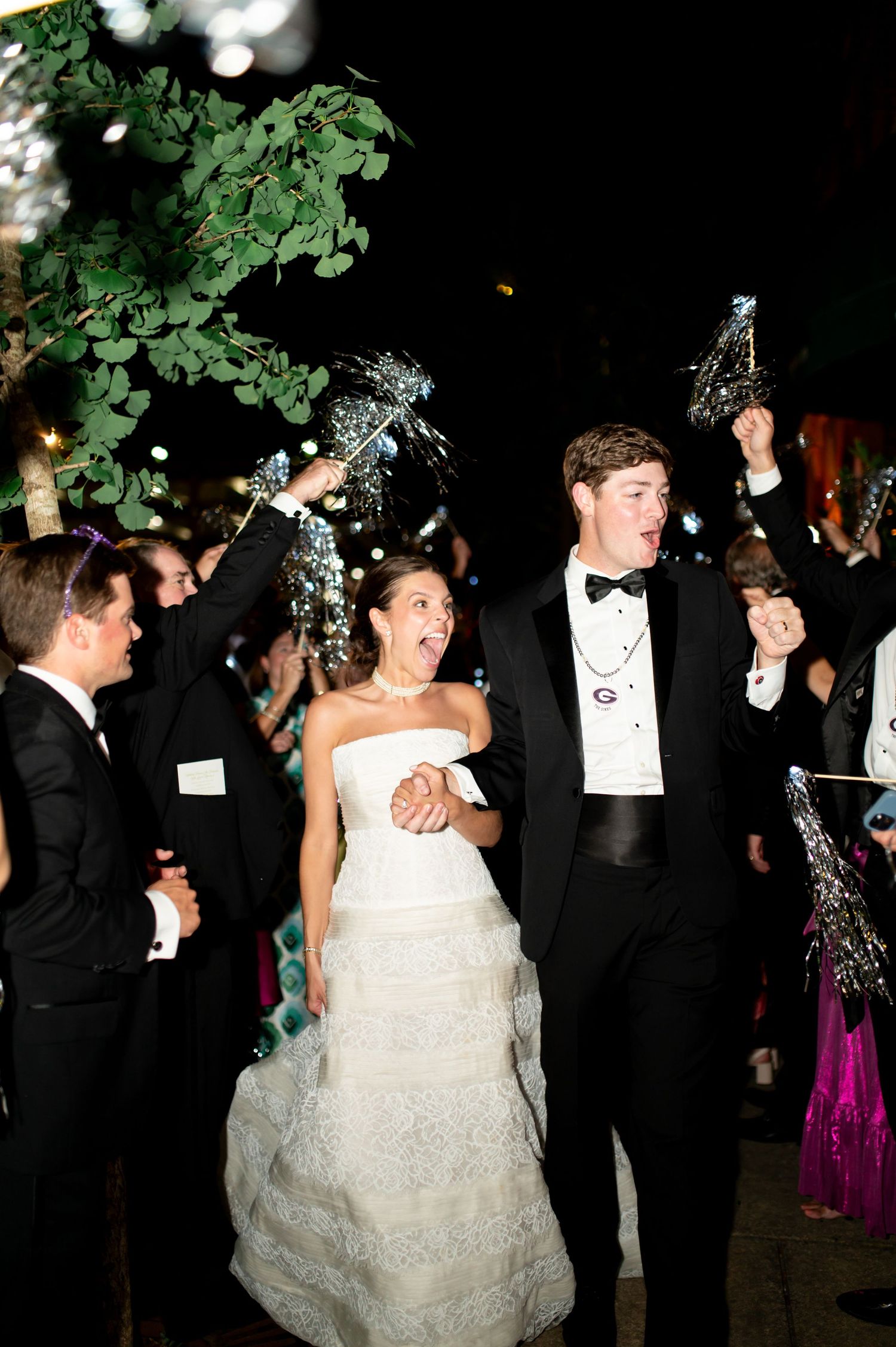 Bride and groom exit wedding reception through cheering guests waving silver sparkle wands at night.