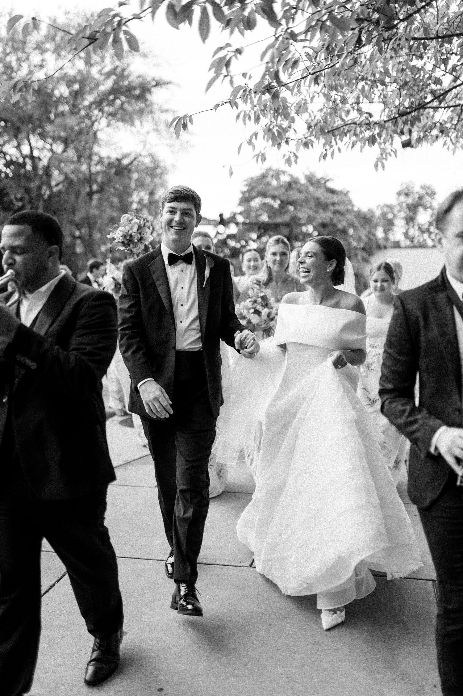 Smiling newlyweds walk hand-in-hand outdoors after wedding ceremony in black and white photo.