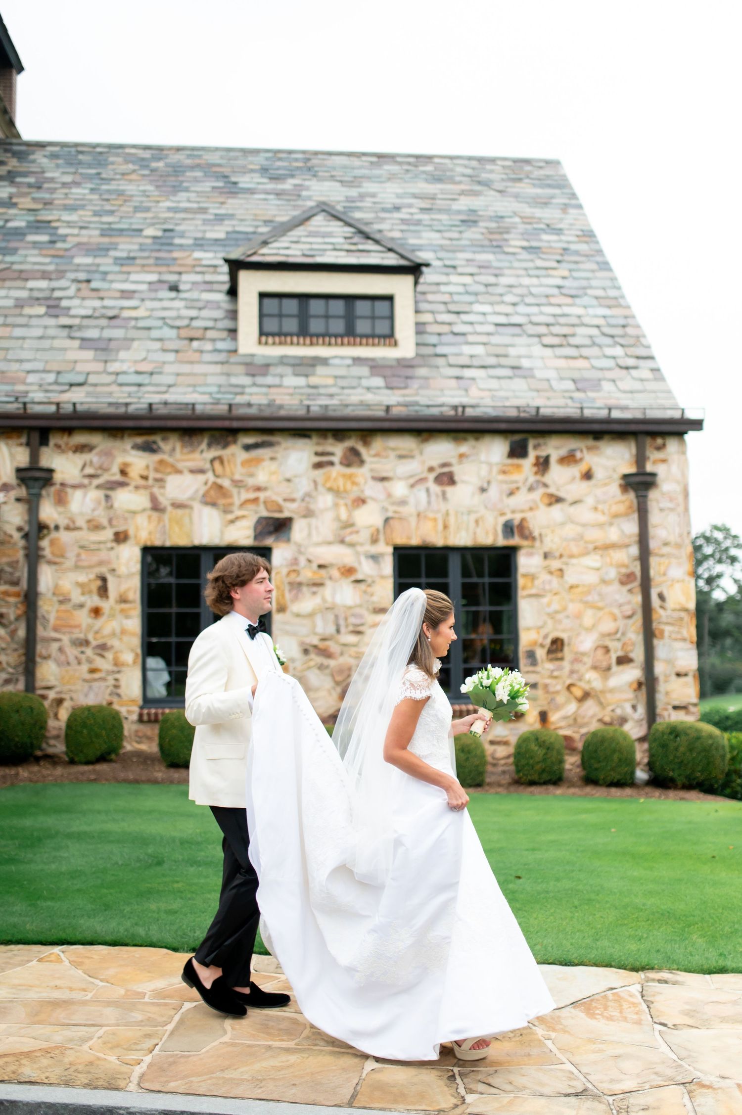 Wedding couple walking outside a rustic stone building with a flowing white dress trailing behind.