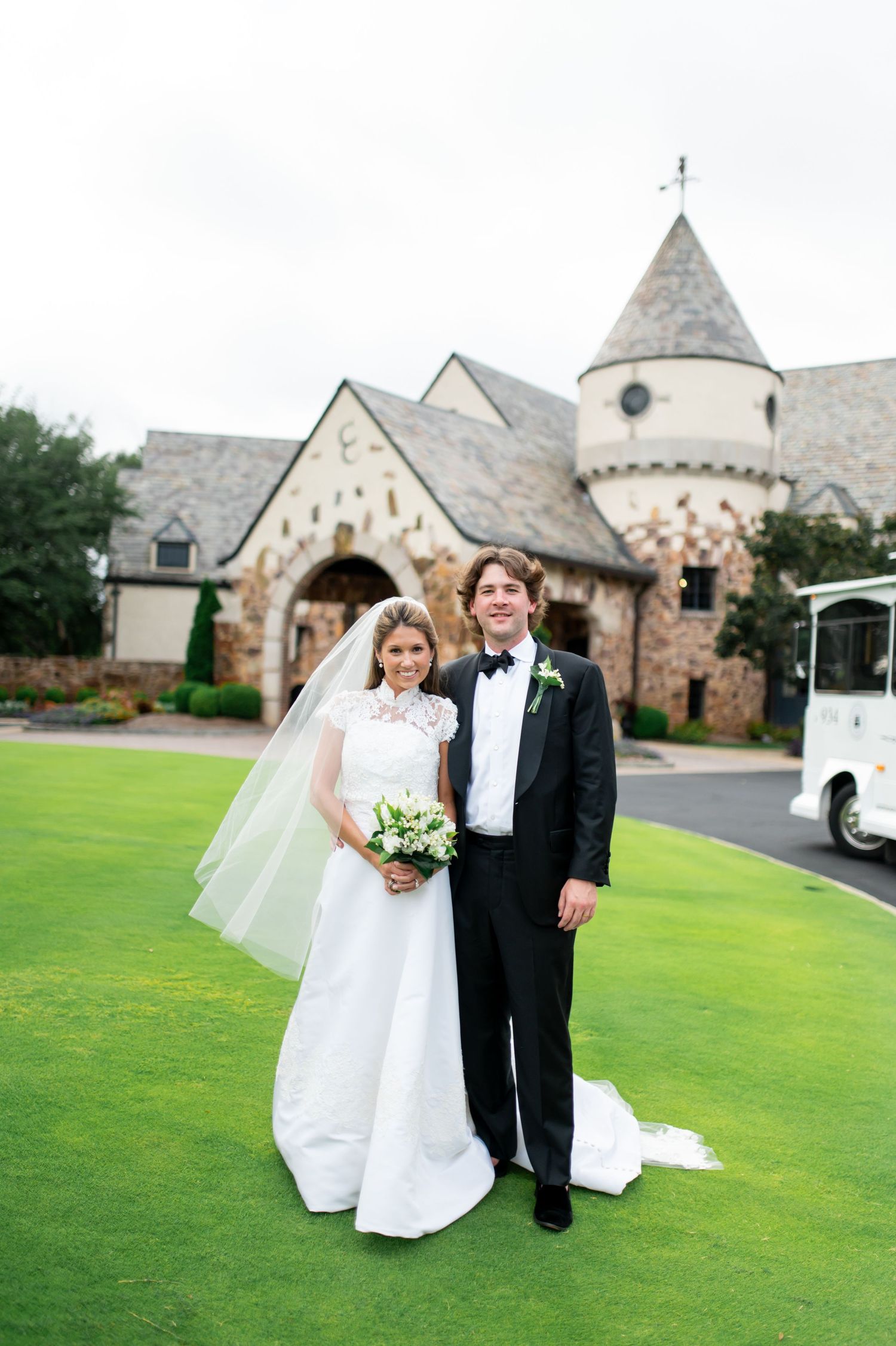 Wedding couple poses on green lawn with stone church architecture in background.