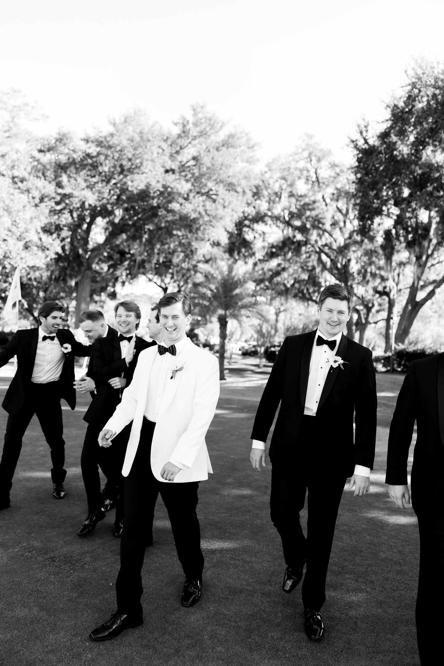 Wedding party members in black tuxedos strike playful poses on a grassy lawn.