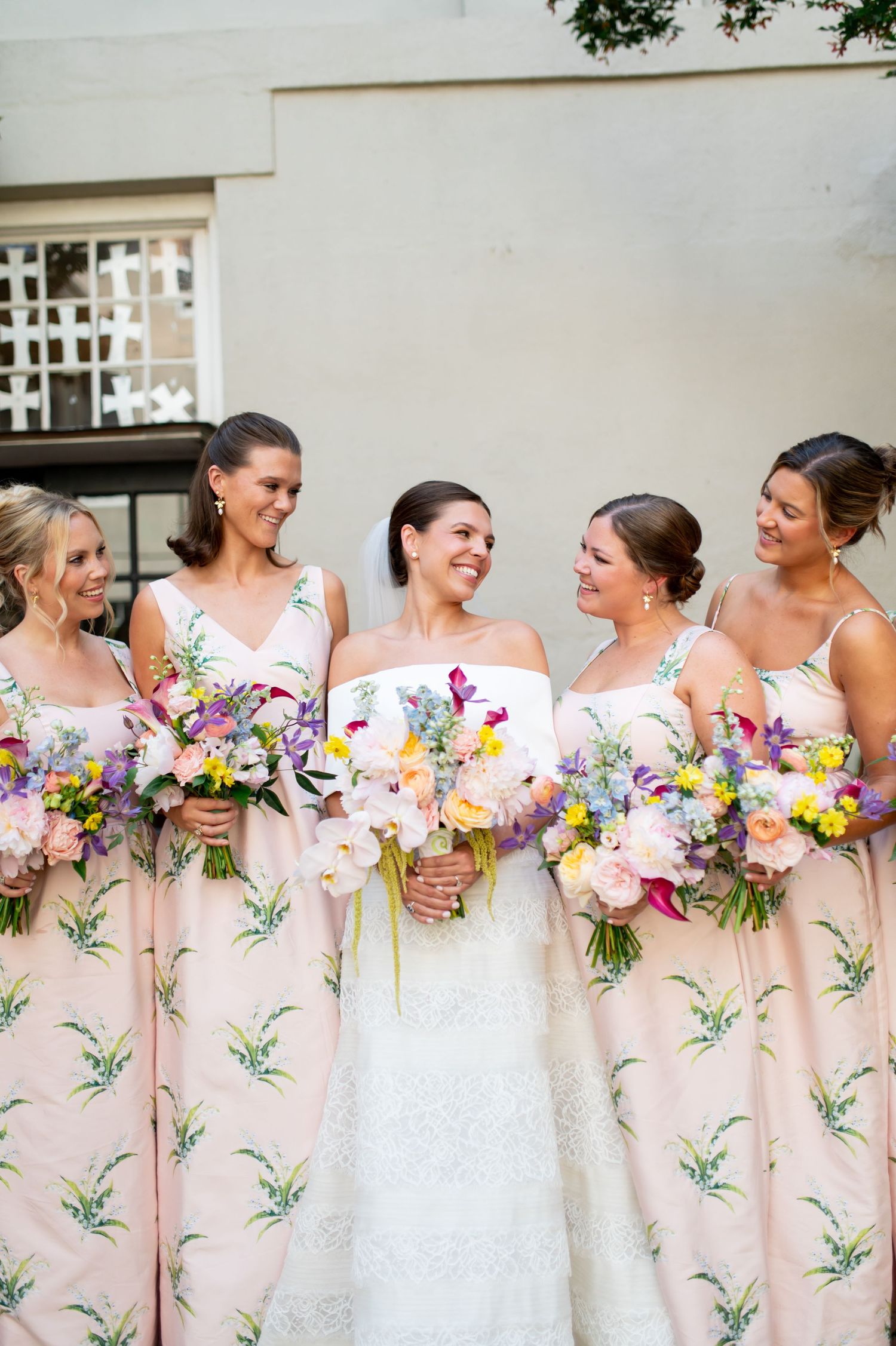 Smiling bride in strapless lace gown poses with four bridesmaids in pink floral dresses holding colorful bouquets.