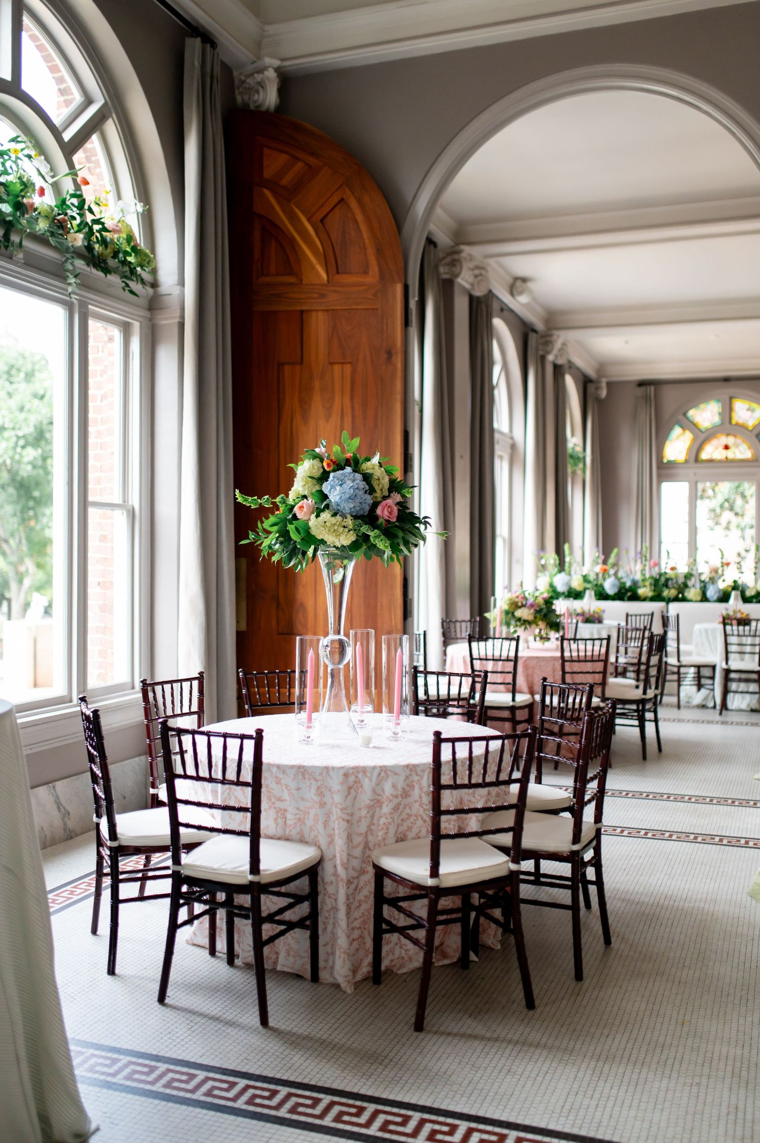Elegant wedding reception hall with tall floral centerpieces, chiavari chairs, and arched windows.