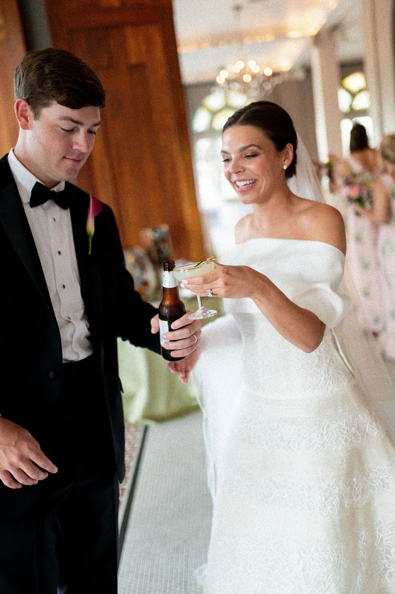 Bride in off-shoulder gown and groom in tuxedo share a toast with cocktails at their elegant wedding reception.