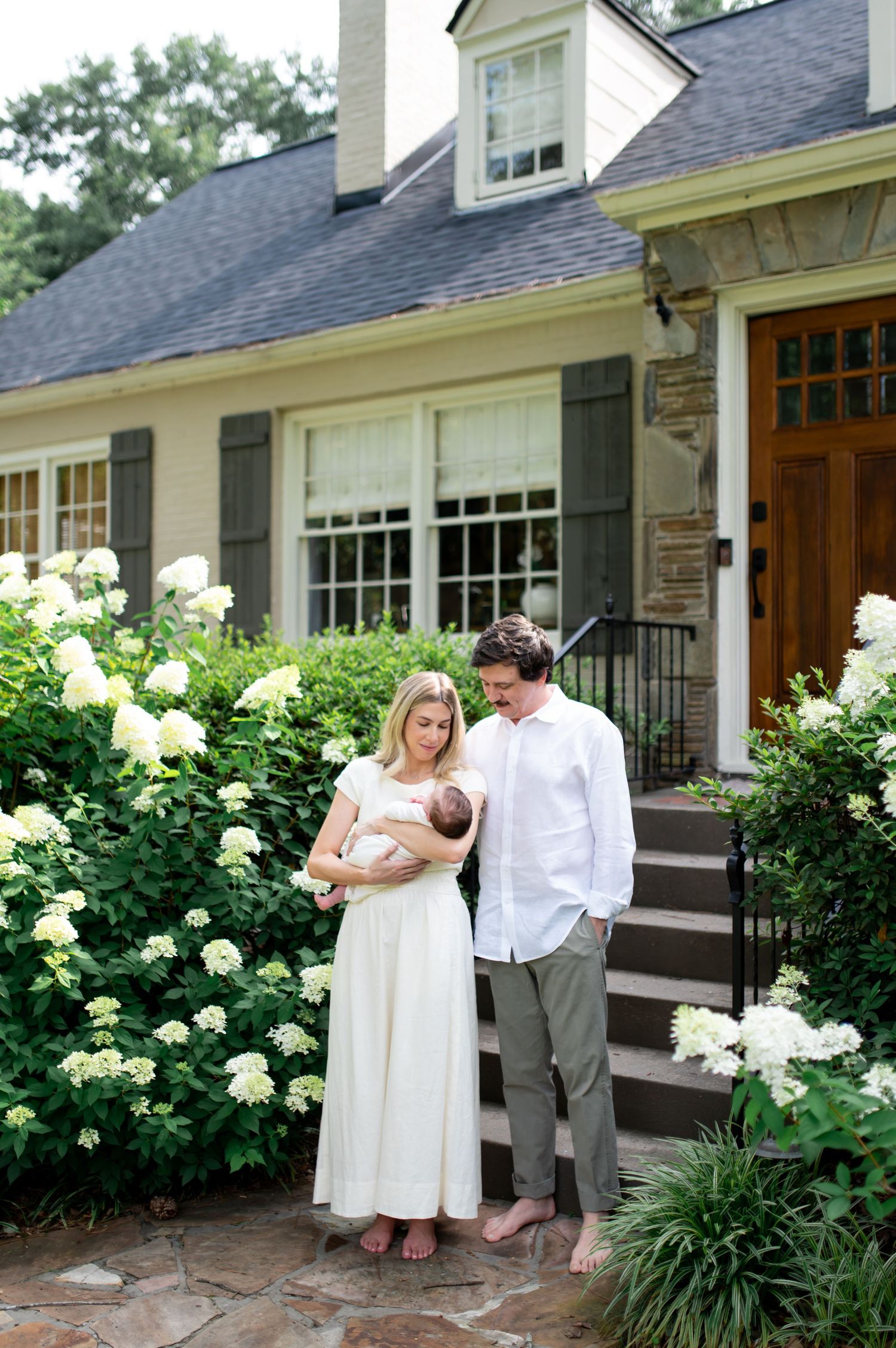 A couple stands outside their charming home holding a newborn baby, surrounded by blooming white hydrangeas.