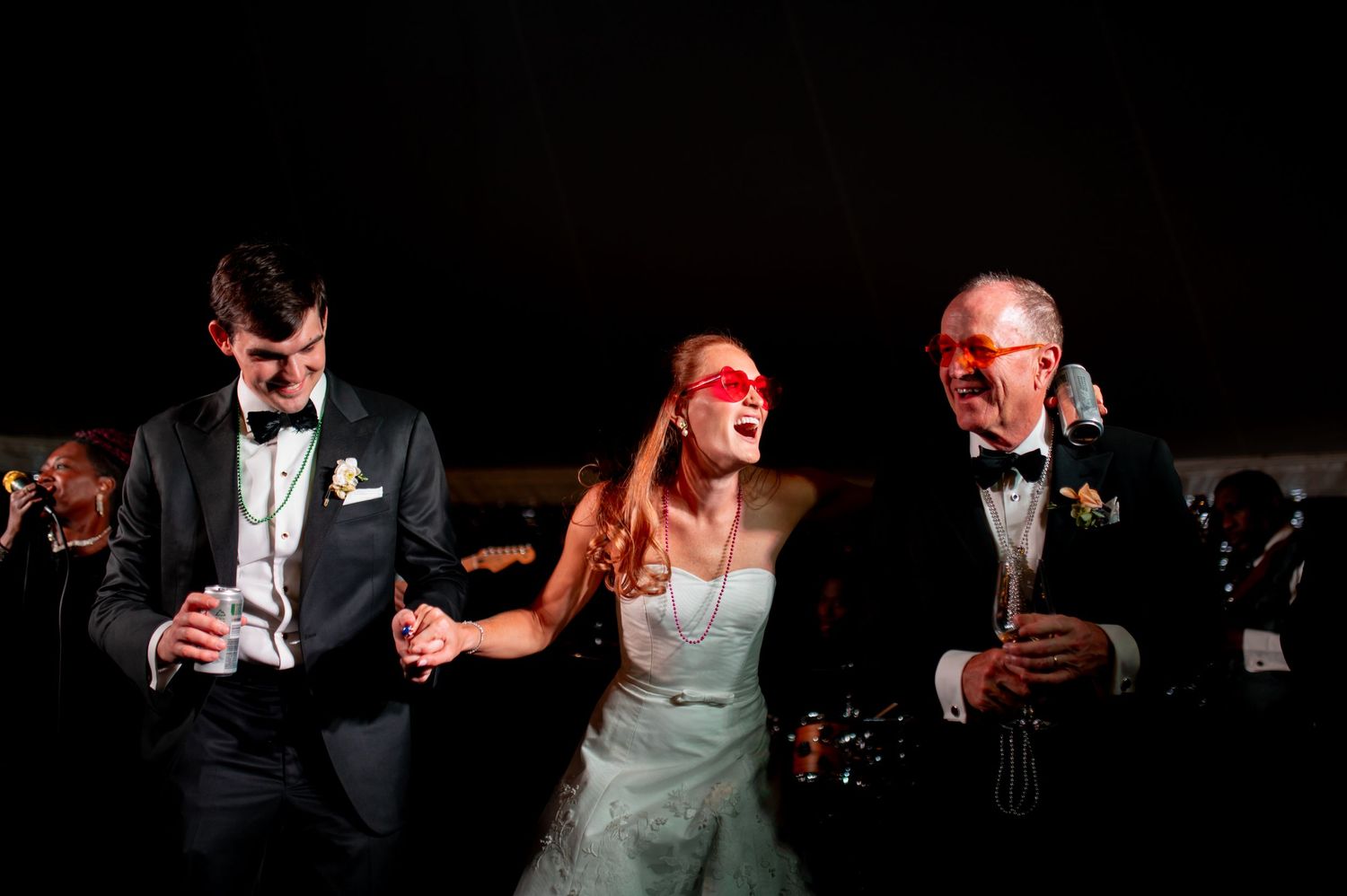 Bride and two guests dance and laugh wearing red masquerade masks at evening wedding reception.