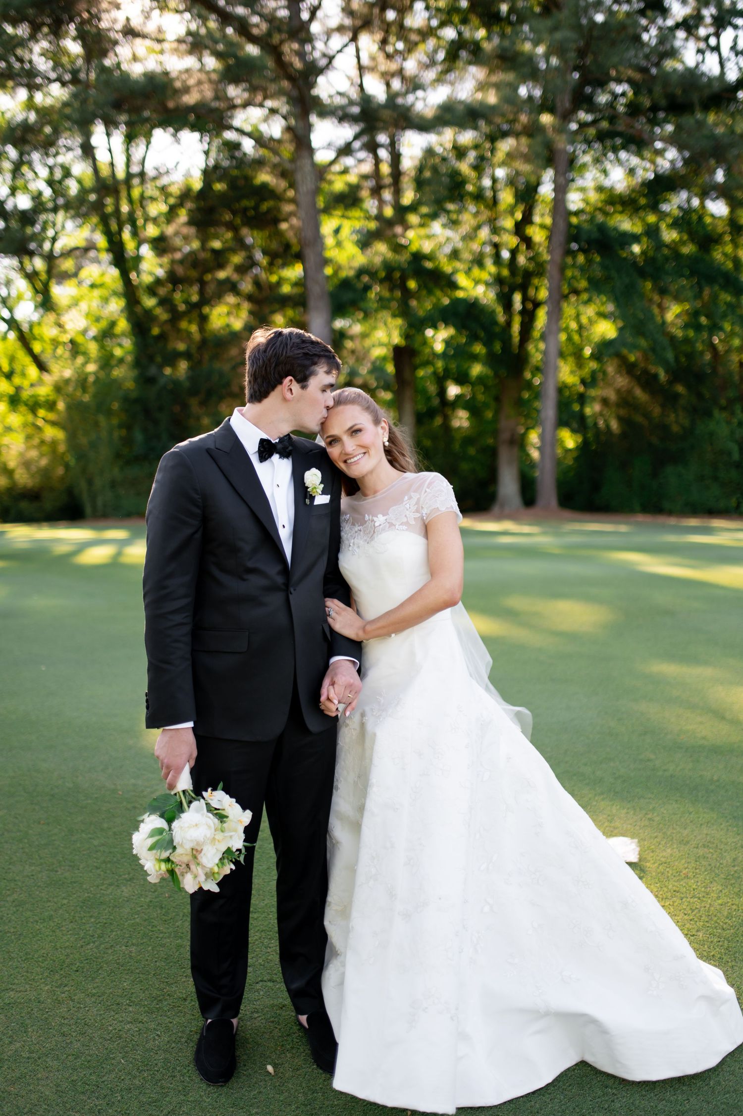 Bride and groom pose on golf course, groom kissing bride's cheek, both holding white flower bouquet.