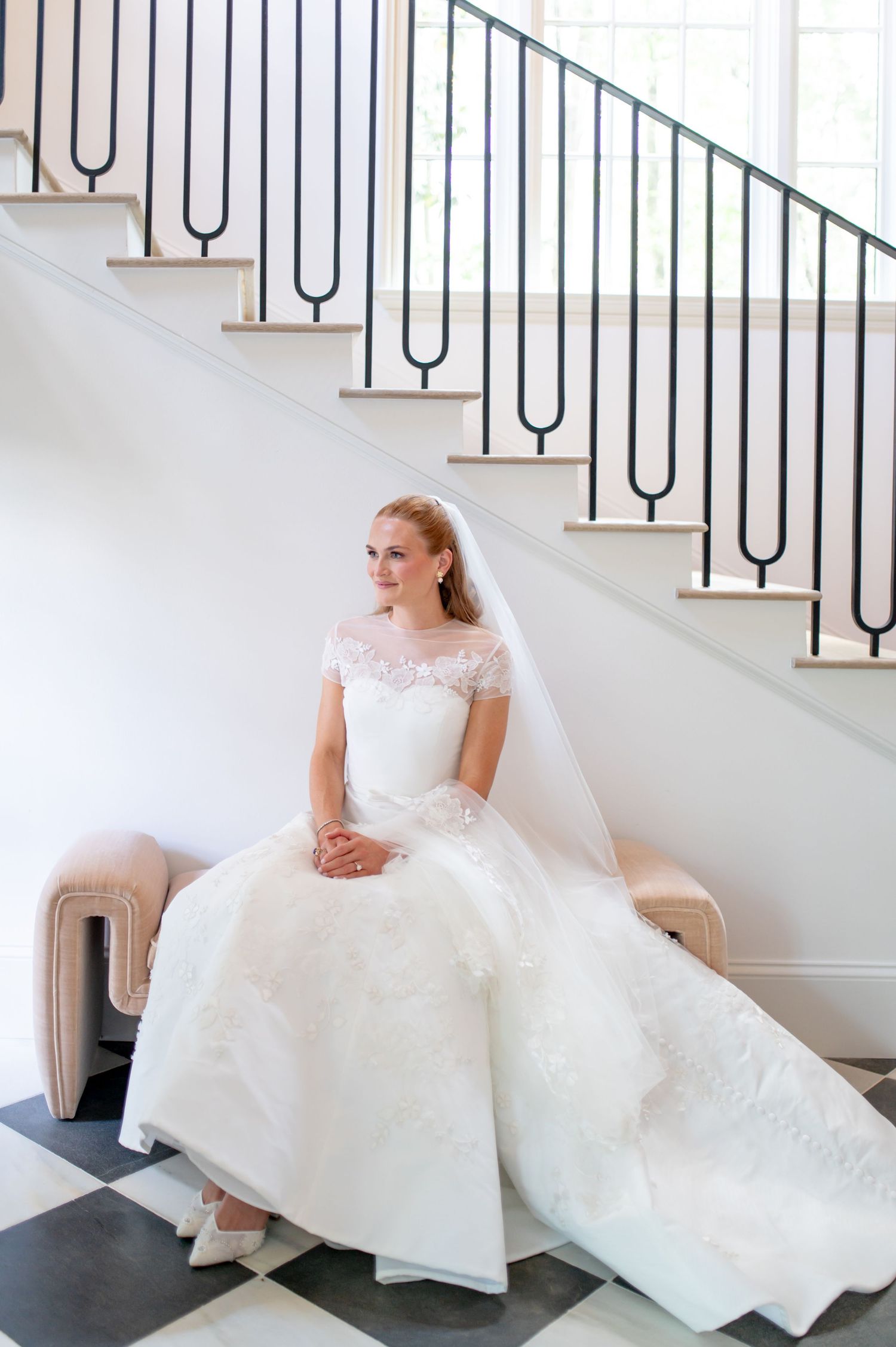 Elegant bride in lace cap-sleeve wedding dress seated by staircase with black iron railing.