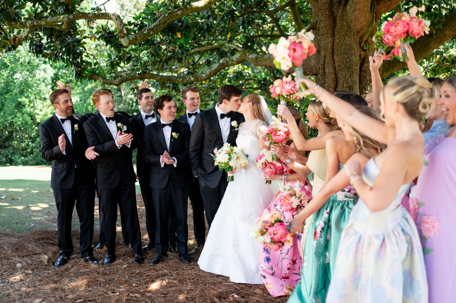 Wedding party celebration under oak tree, bridesmaids raising pink bouquets, groomsmen in black tuxedos.