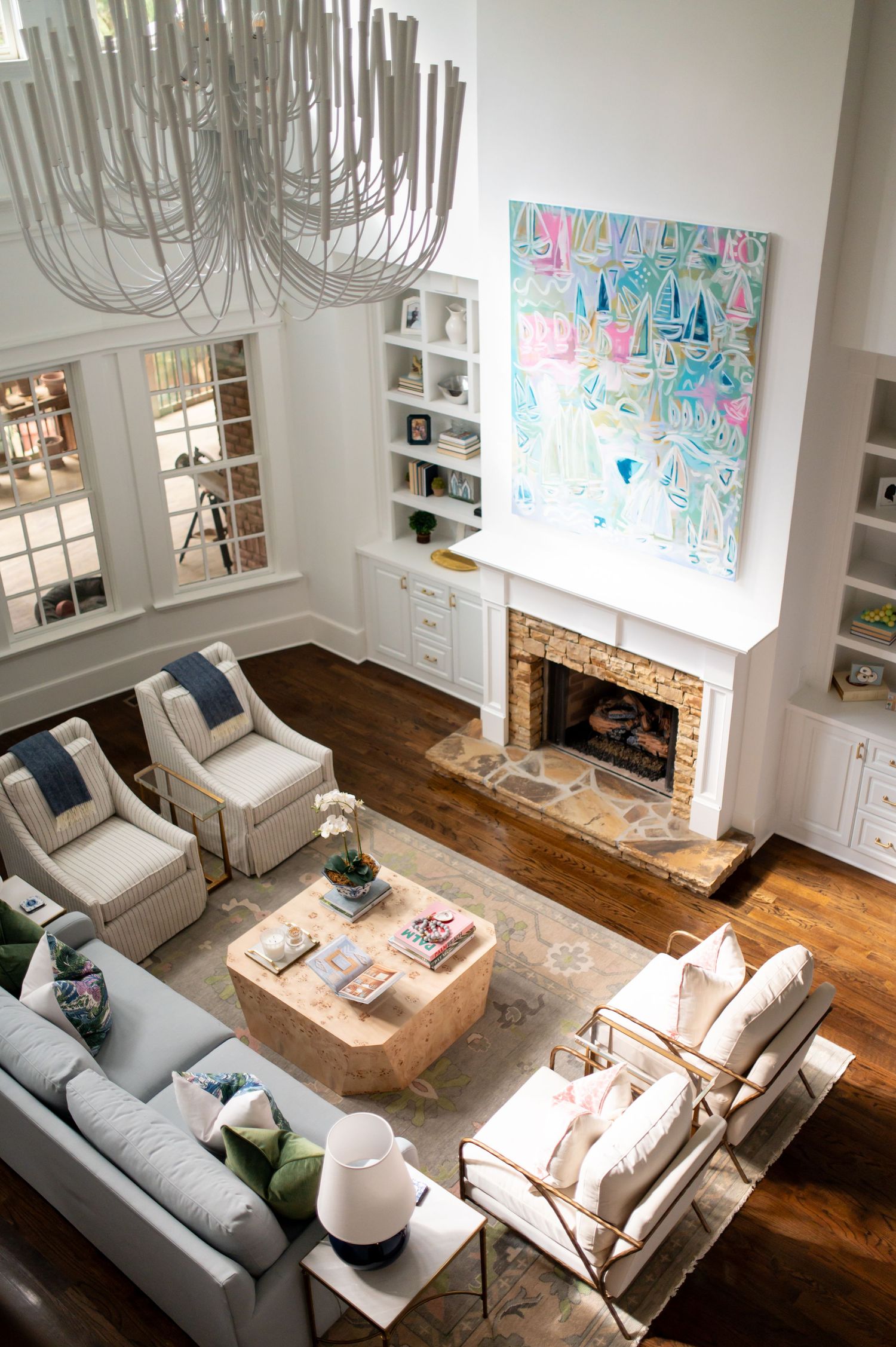 Modern coastal living room with high ceiling, chandelier, stone fireplace, built-in shelving and bright abstract artwork.