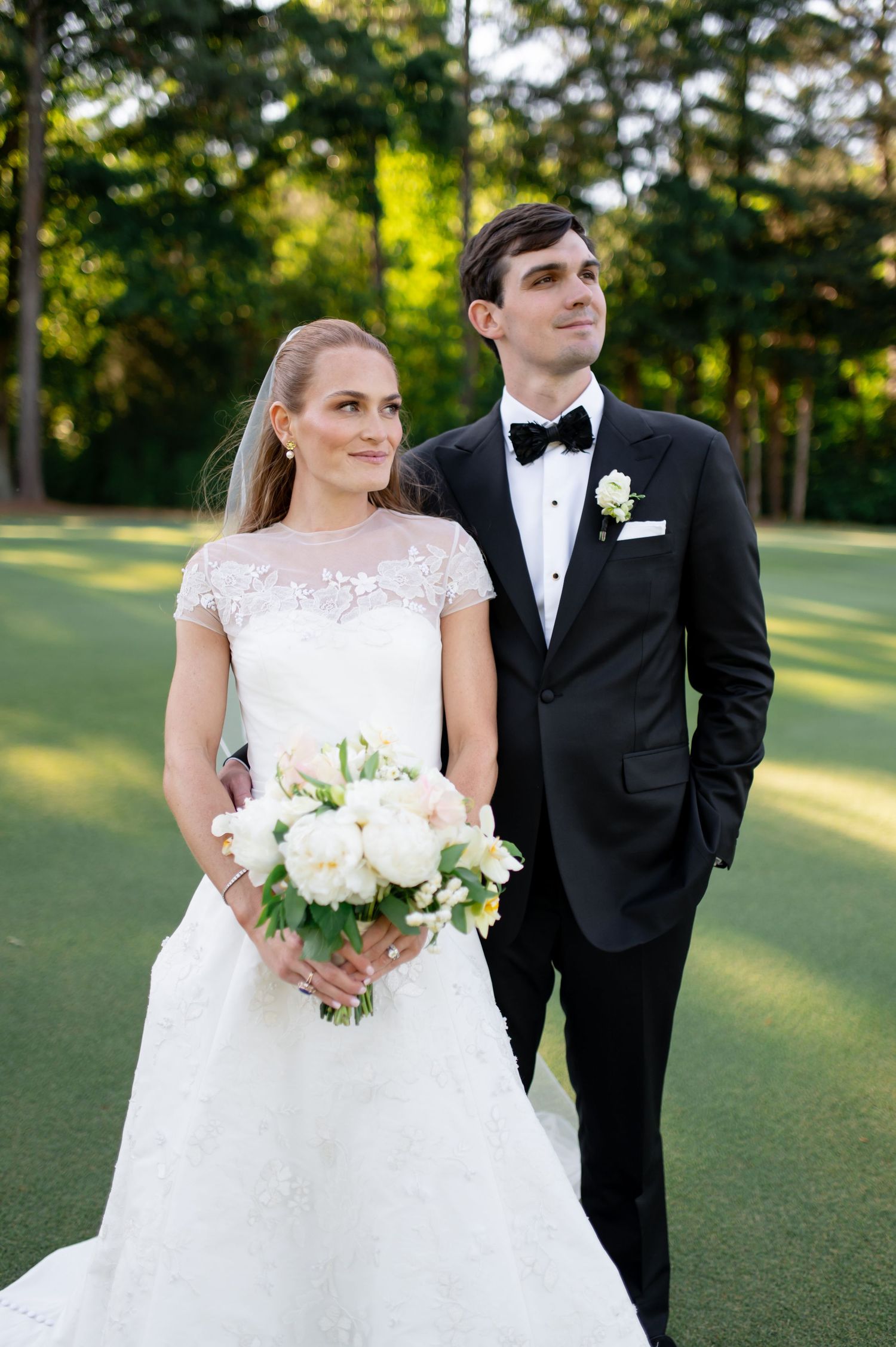 Bride in lace cap sleeve gown holding peony bouquet stands with groom in black tuxedo.