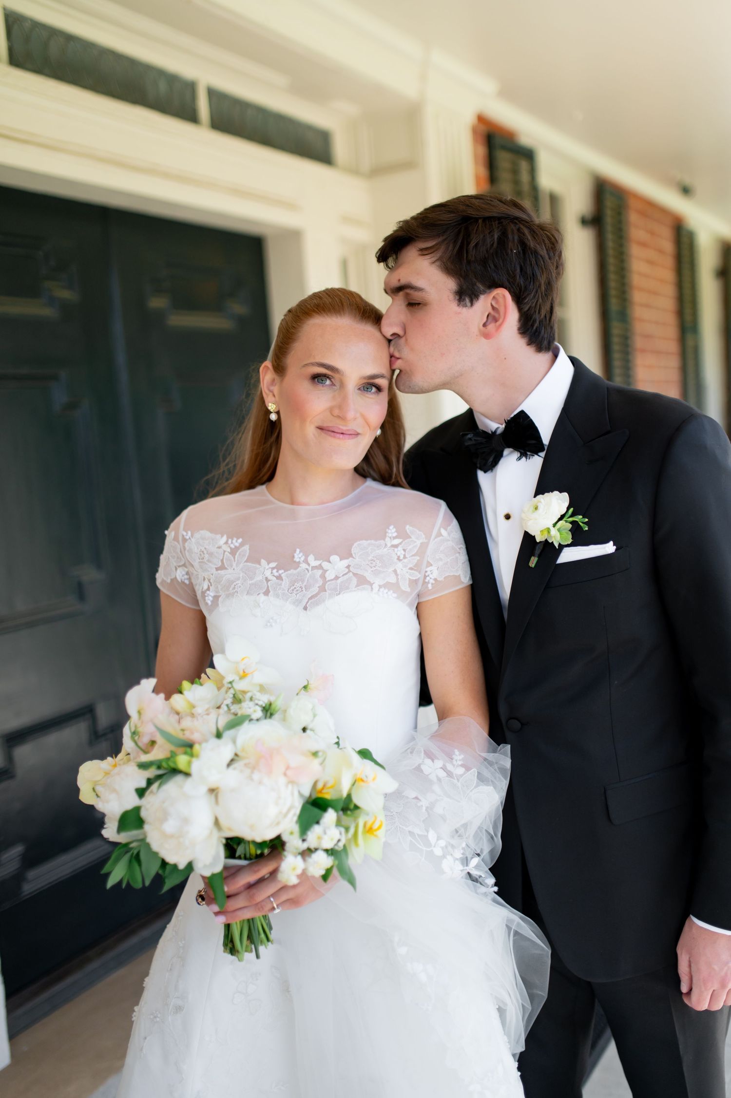 Groom in black tuxedo kisses bride on cheek as she holds white peony bouquet.