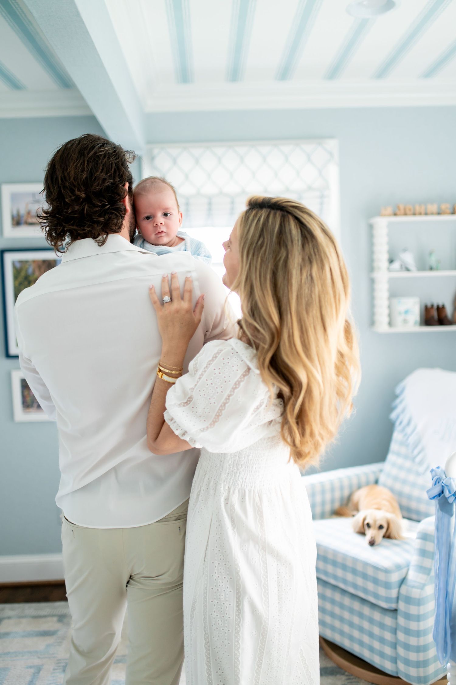 Parents holding newborn baby in a soft blue nursery room with gingham chair and a golden dog nearby.