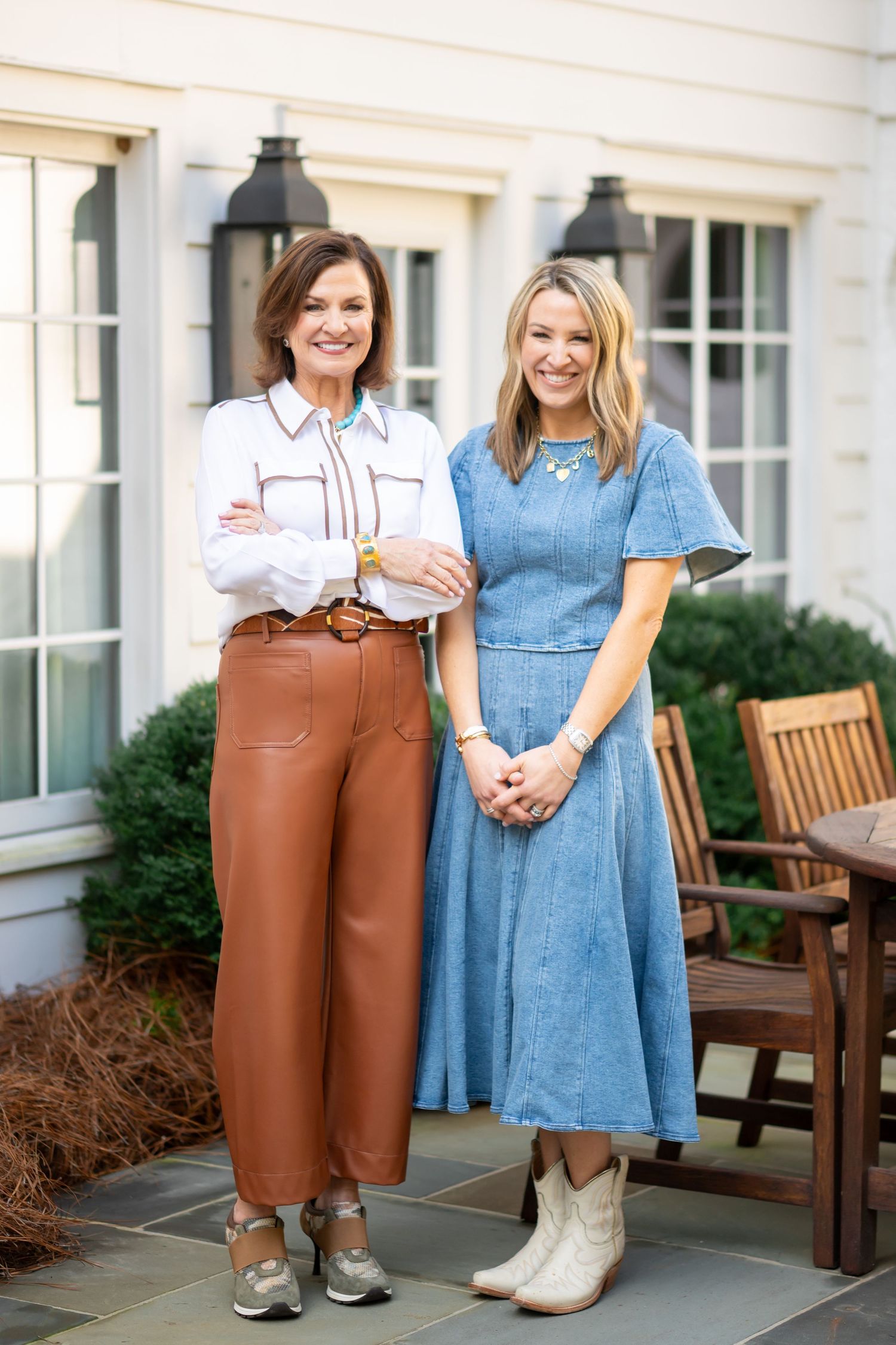Two women smiling outdoors, one in brown leather pants and white shirt, other in blue denim midi dress with white boots.