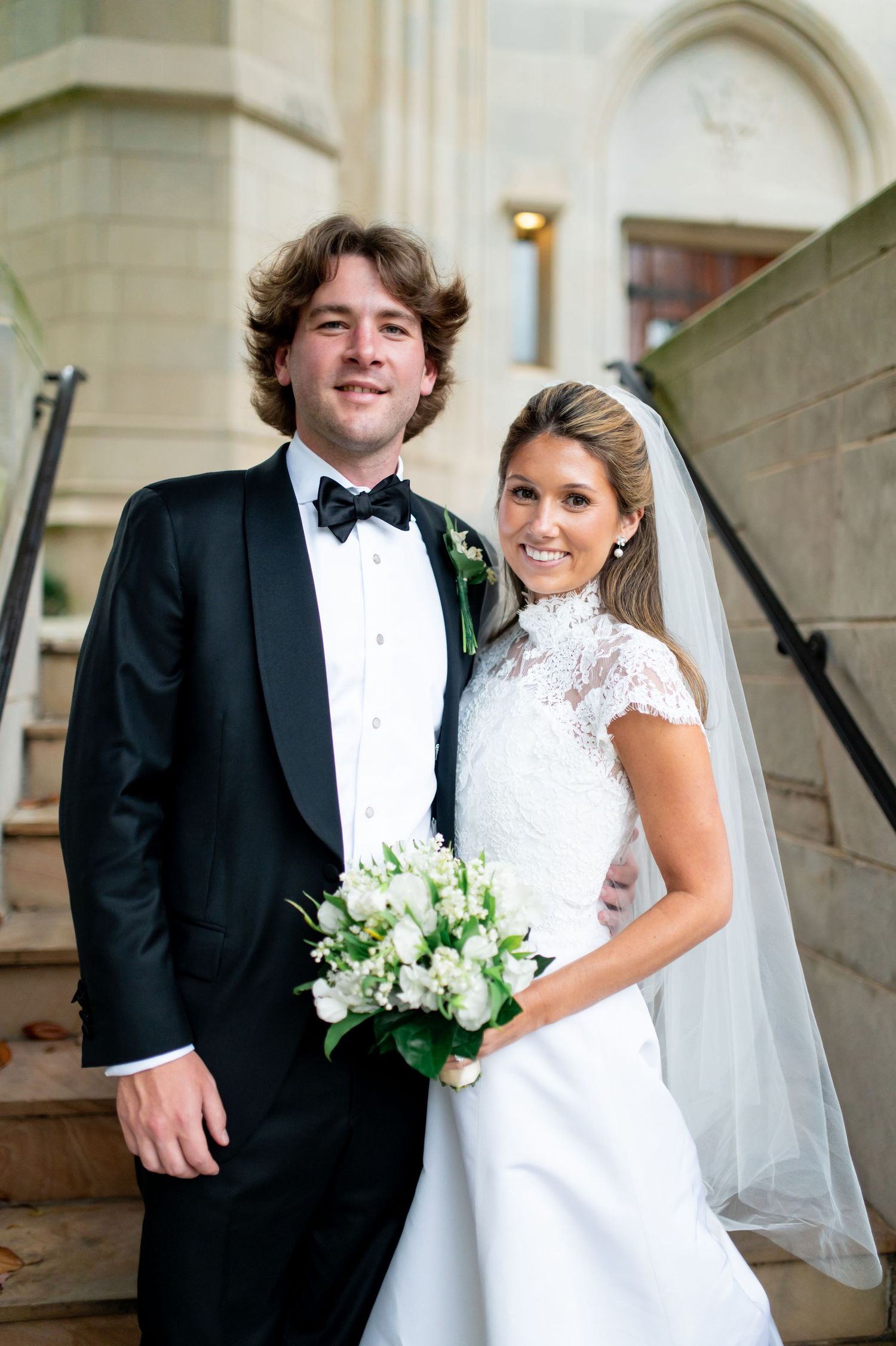A bride and groom pose together on church steps in formal wedding attire.