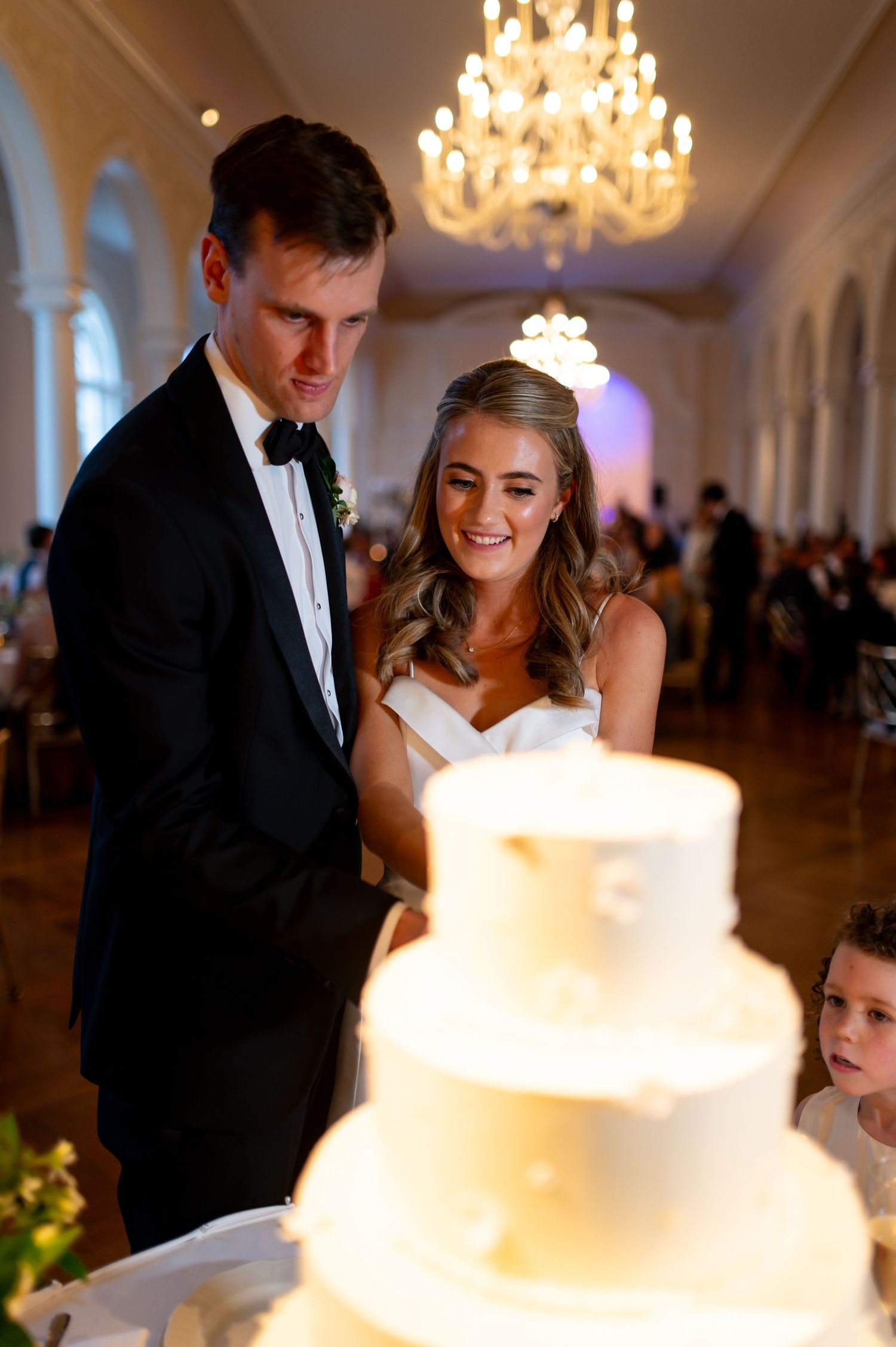 Wedding cake cutting ceremony in an elegant ballroom with crystal chandeliers overhead.