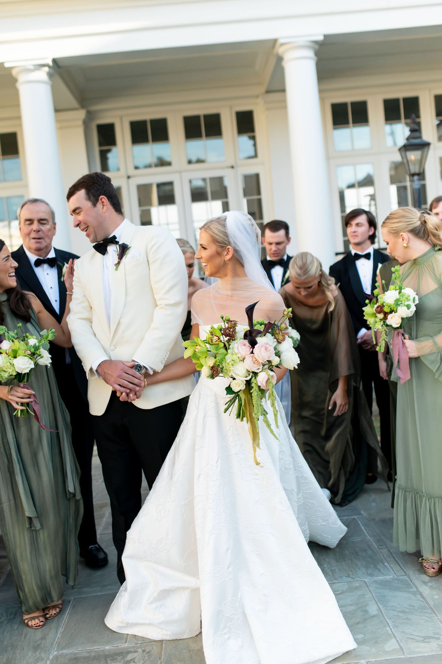 Wedding party poses on a columned porch with bridesmaids in sage green dresses.