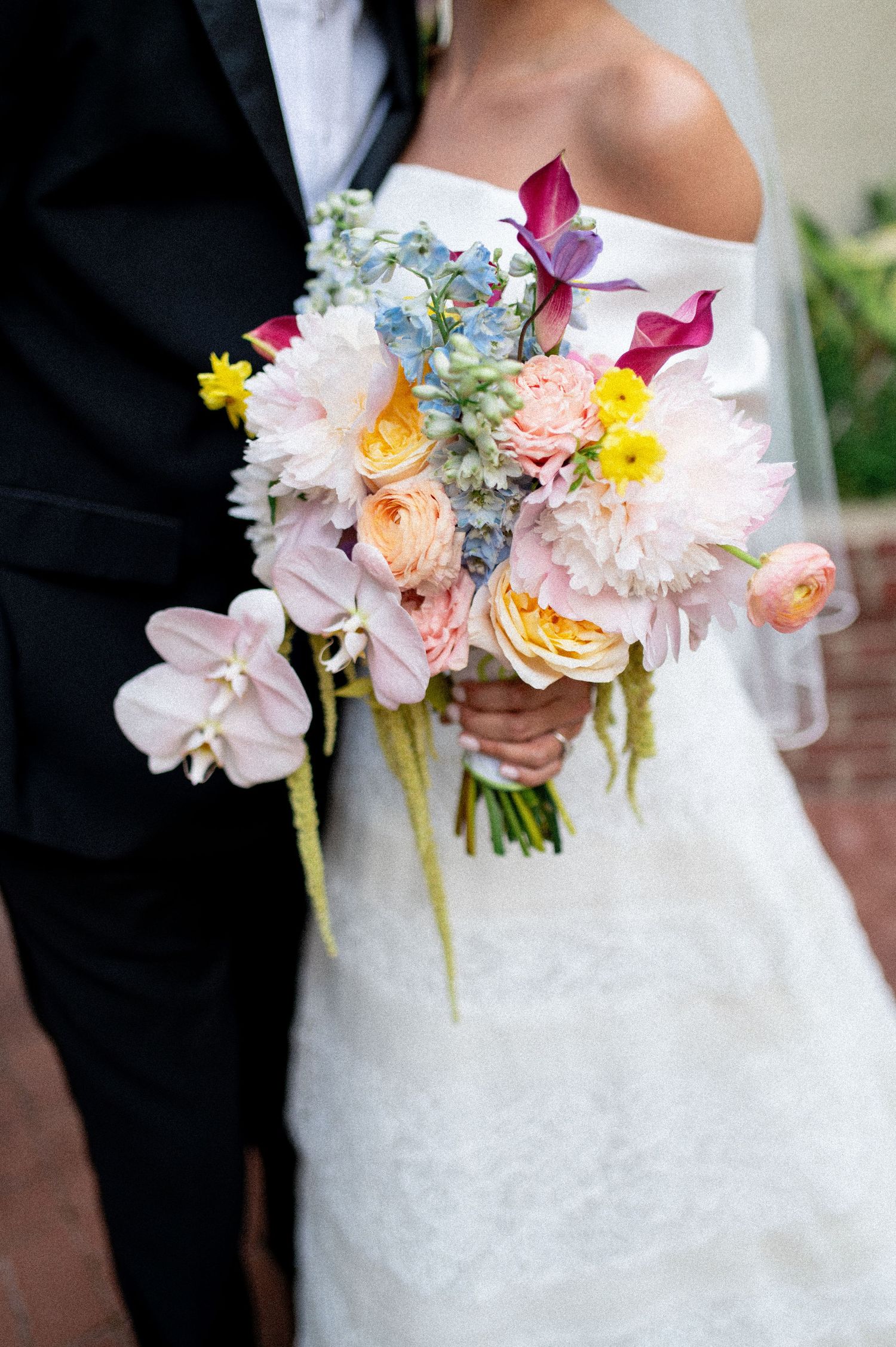Bride holding a colorful wedding bouquet with pink dahlias, peach roses, blue flowers, and orchids in white lace gown.