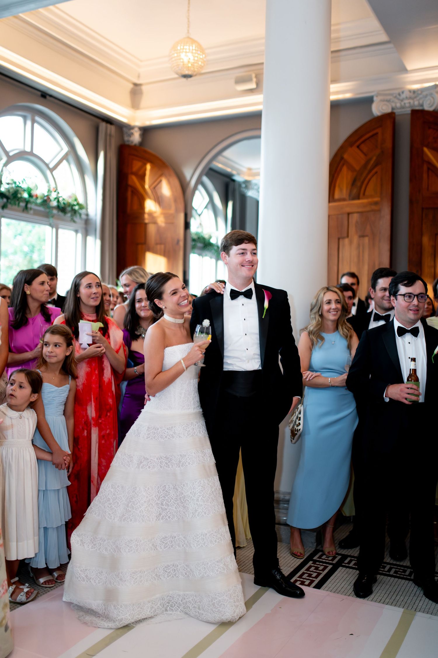 Bride and groom stand together smiling at wedding reception surrounded by guests in elegant white columned ballroom.