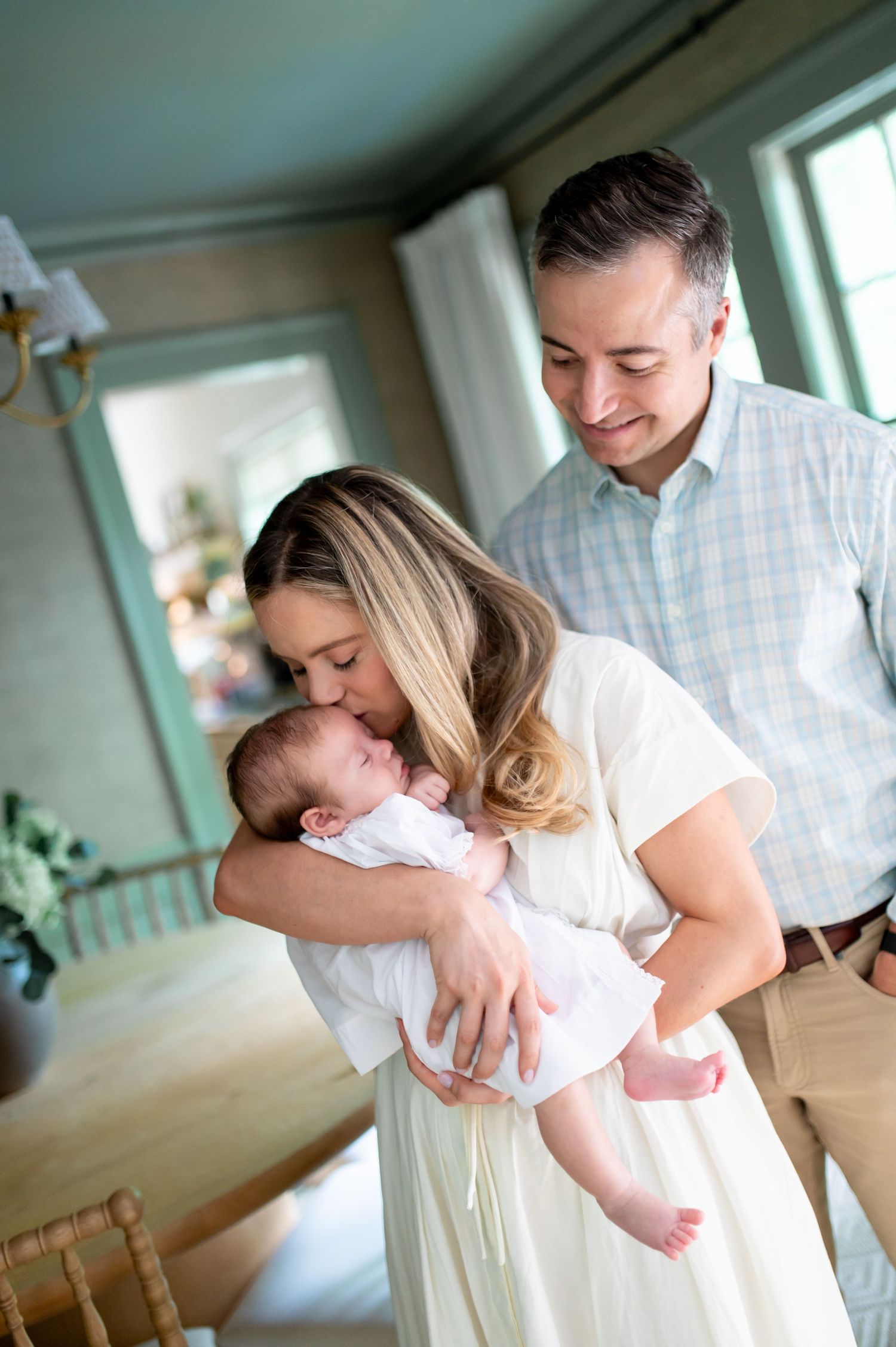 A mother tenderly kisses her newborn baby while the proud father smiles warmly in a bright, elegant home setting.