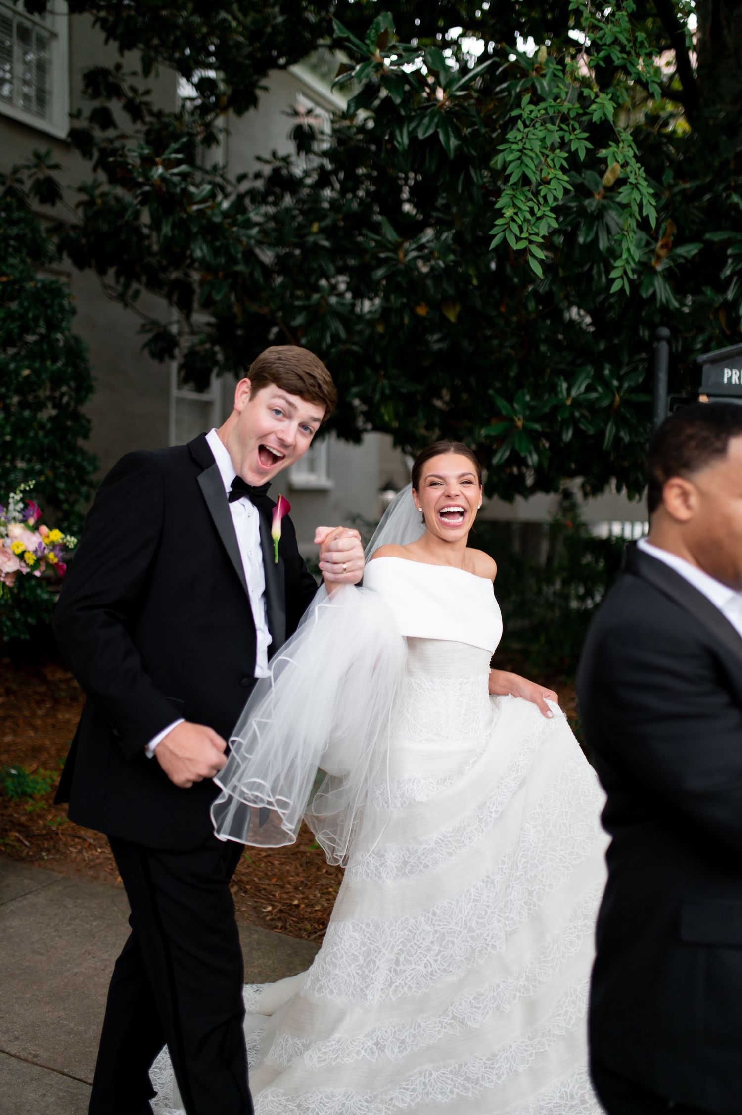 Joyful bride and groom laugh as groom lifts bride's lace gown while walking outdoors after wedding ceremony.