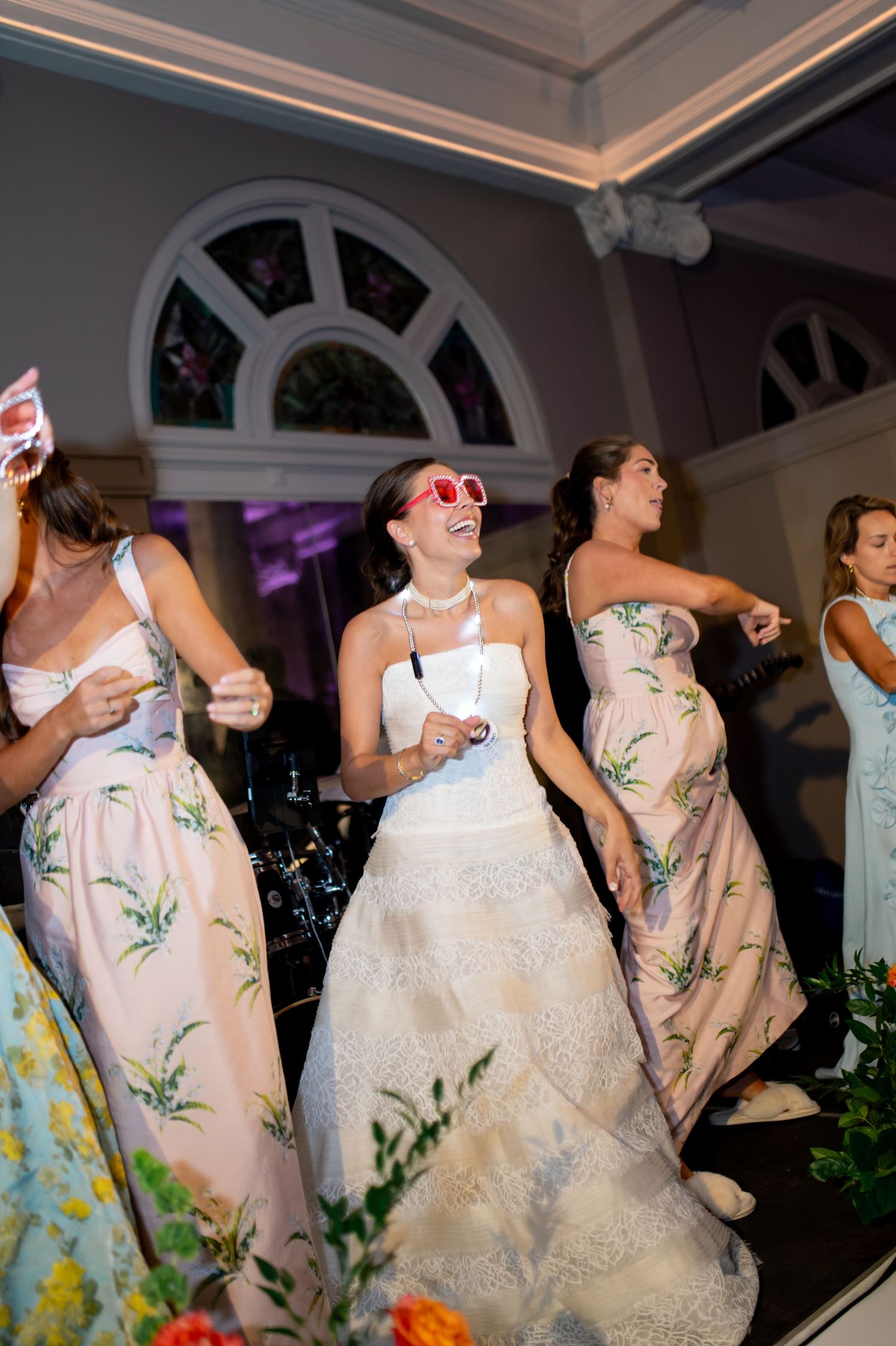 Bride wearing fun pink sunglasses dances joyfully with bridesmaids in floral gowns at lively wedding reception.