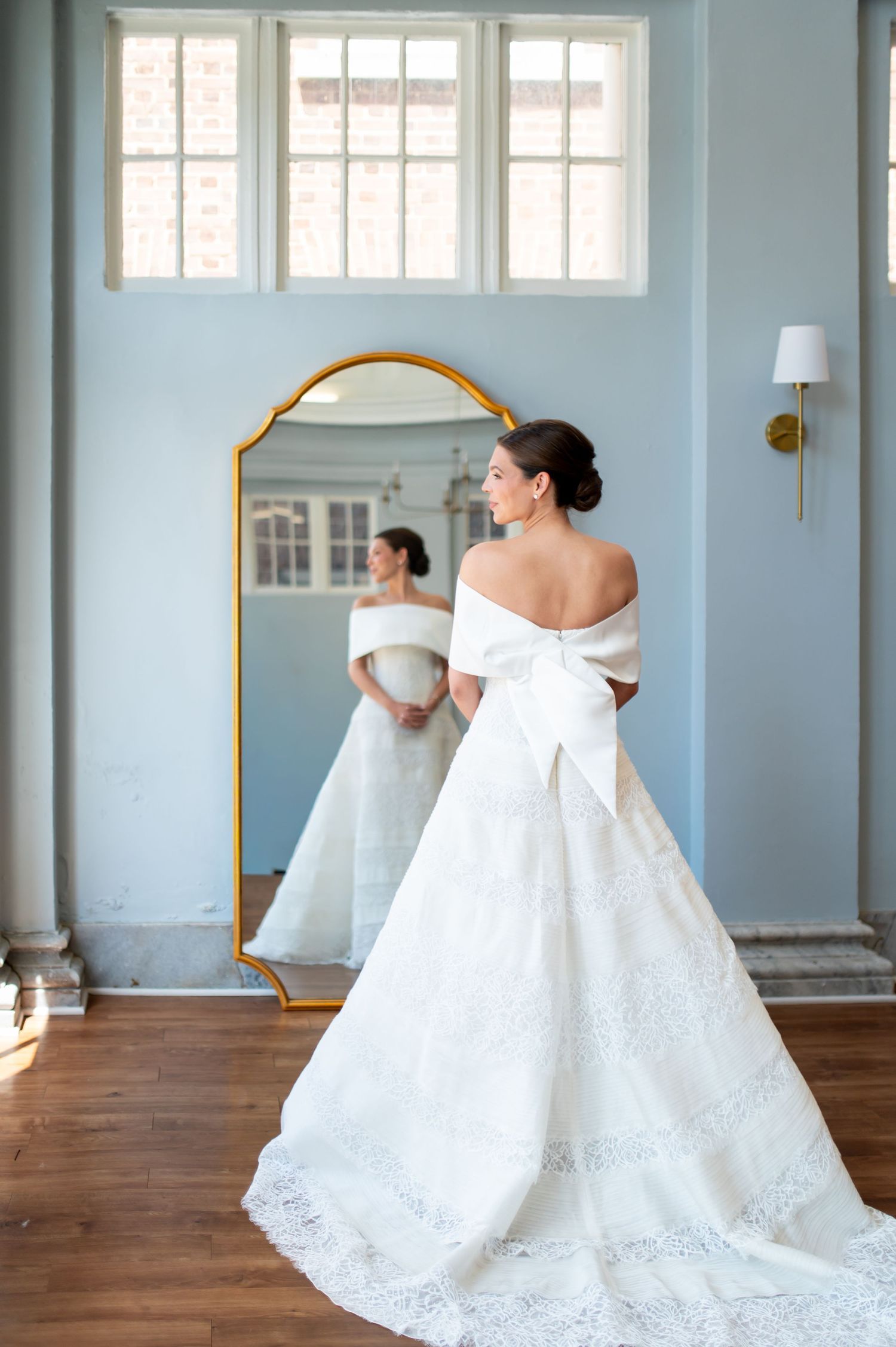 Bride in off-shoulder white ballgown with large bow, standing before gold-framed mirror in blue room.