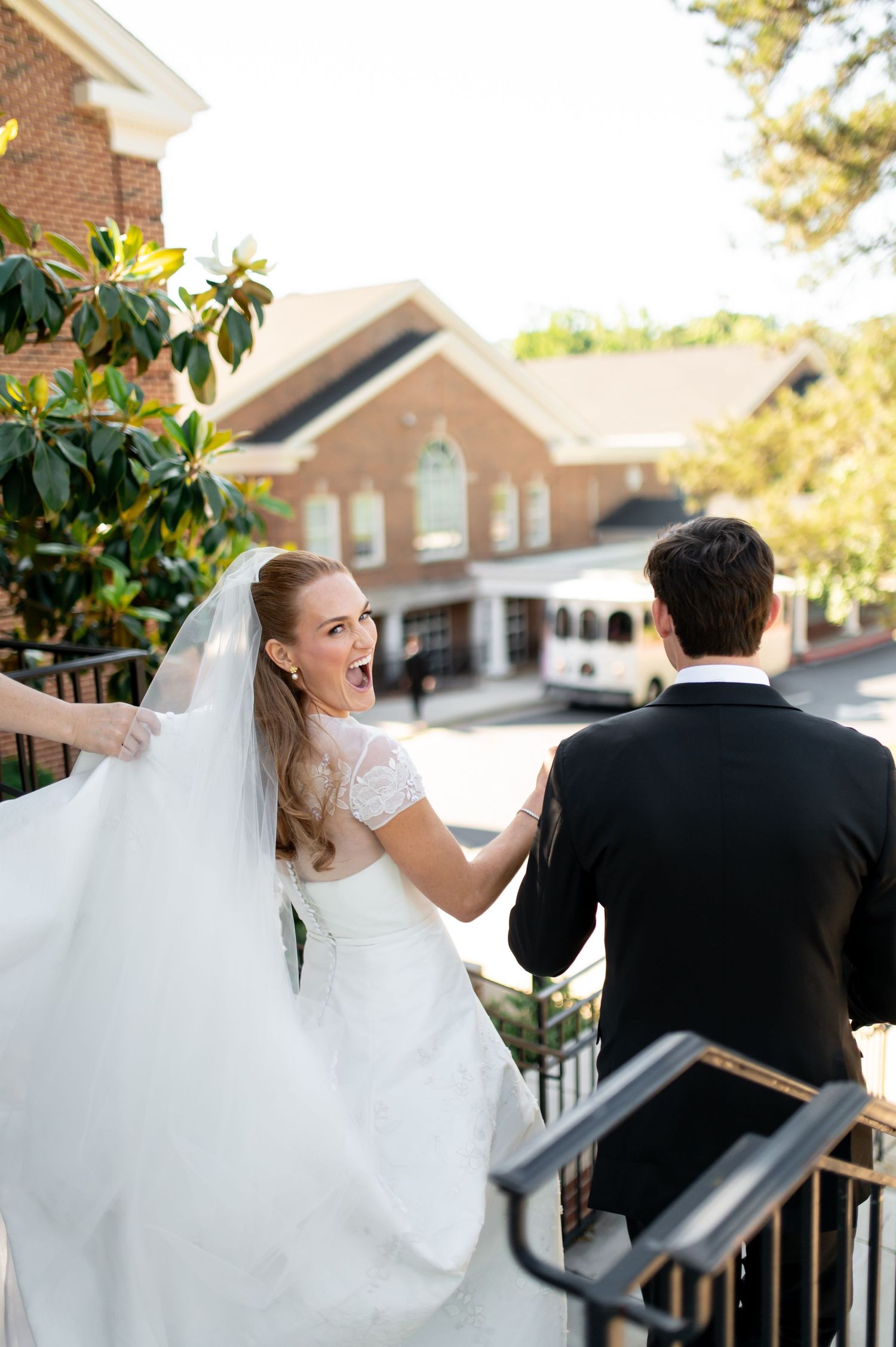 Excited bride in lace gown looks back while ascending stairs with groom outside church.
