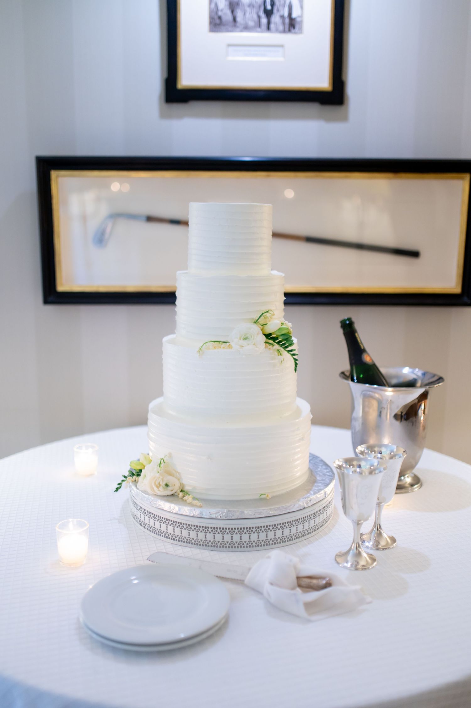 Elegant four-tier white ribbed wedding cake with floral accents on silver stand with champagne bucket.