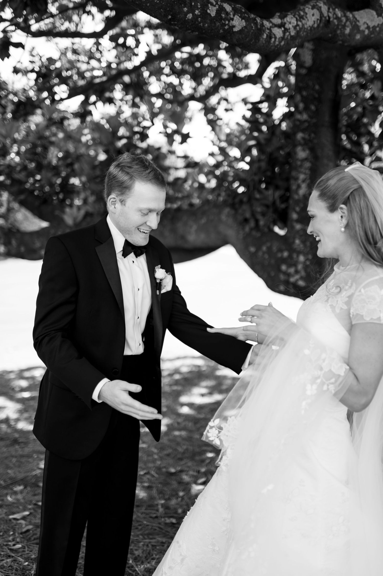 Groom in black tuxedo reacts with joy seeing bride in lace wedding gown during outdoor first look ceremony.