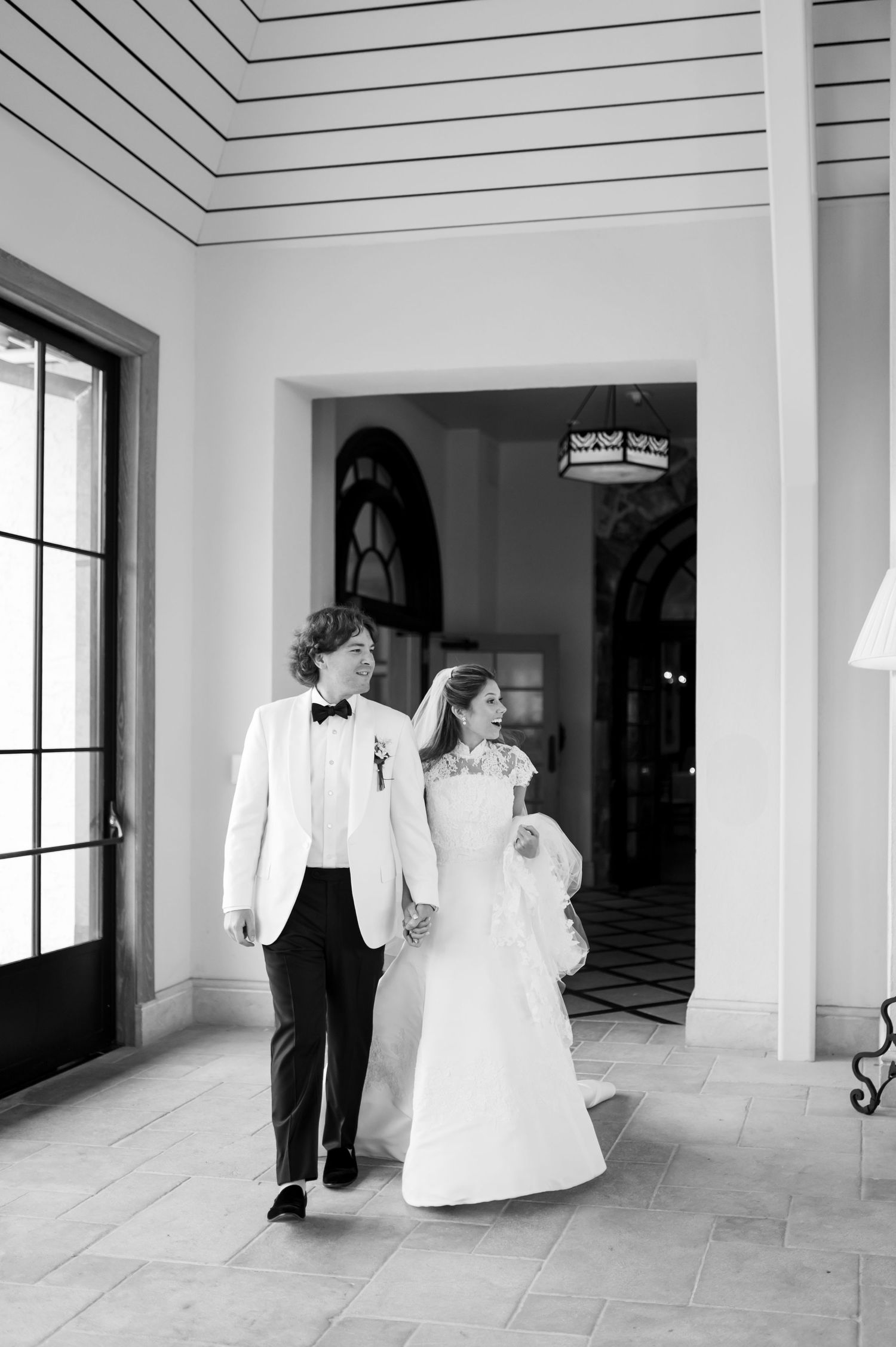 Black and white photo of couple walking through modern hallway with large windows.