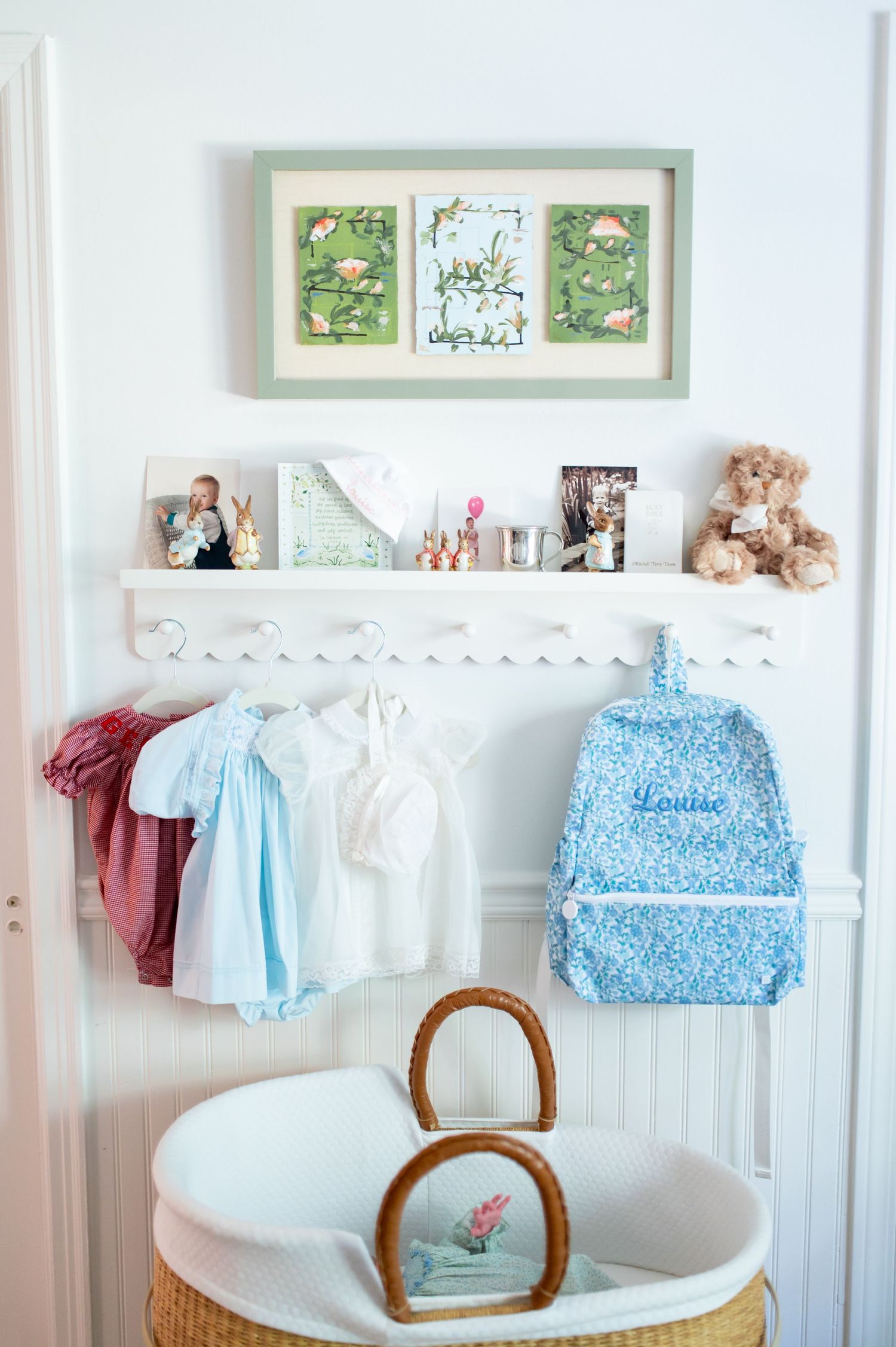 A nursery wall with framed artwork, white shelf displaying photos and toys, hanging children's clothes, and a wicker basket below.