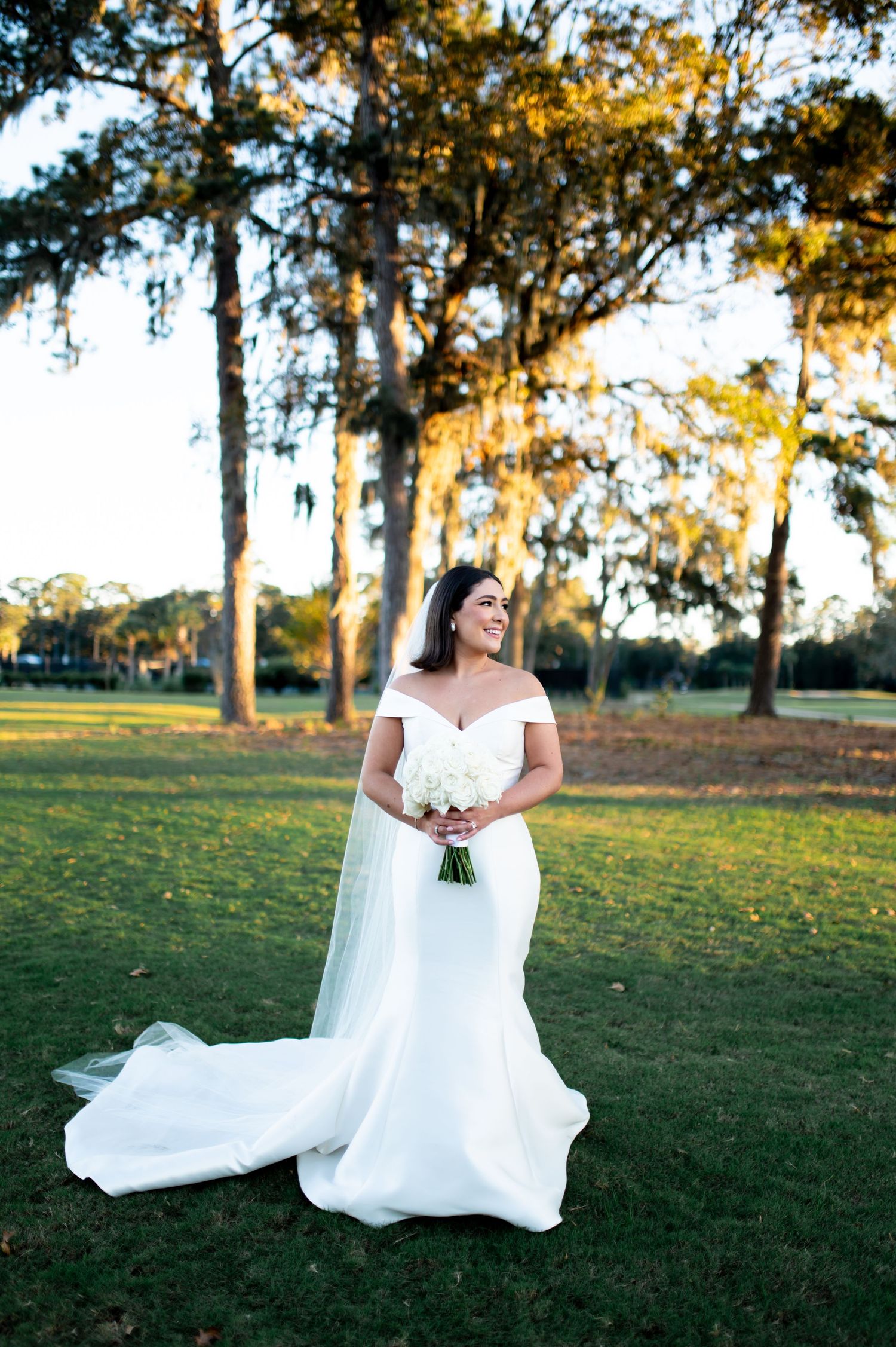 Bride in elegant white gown poses on grassy lawn during golden hour.