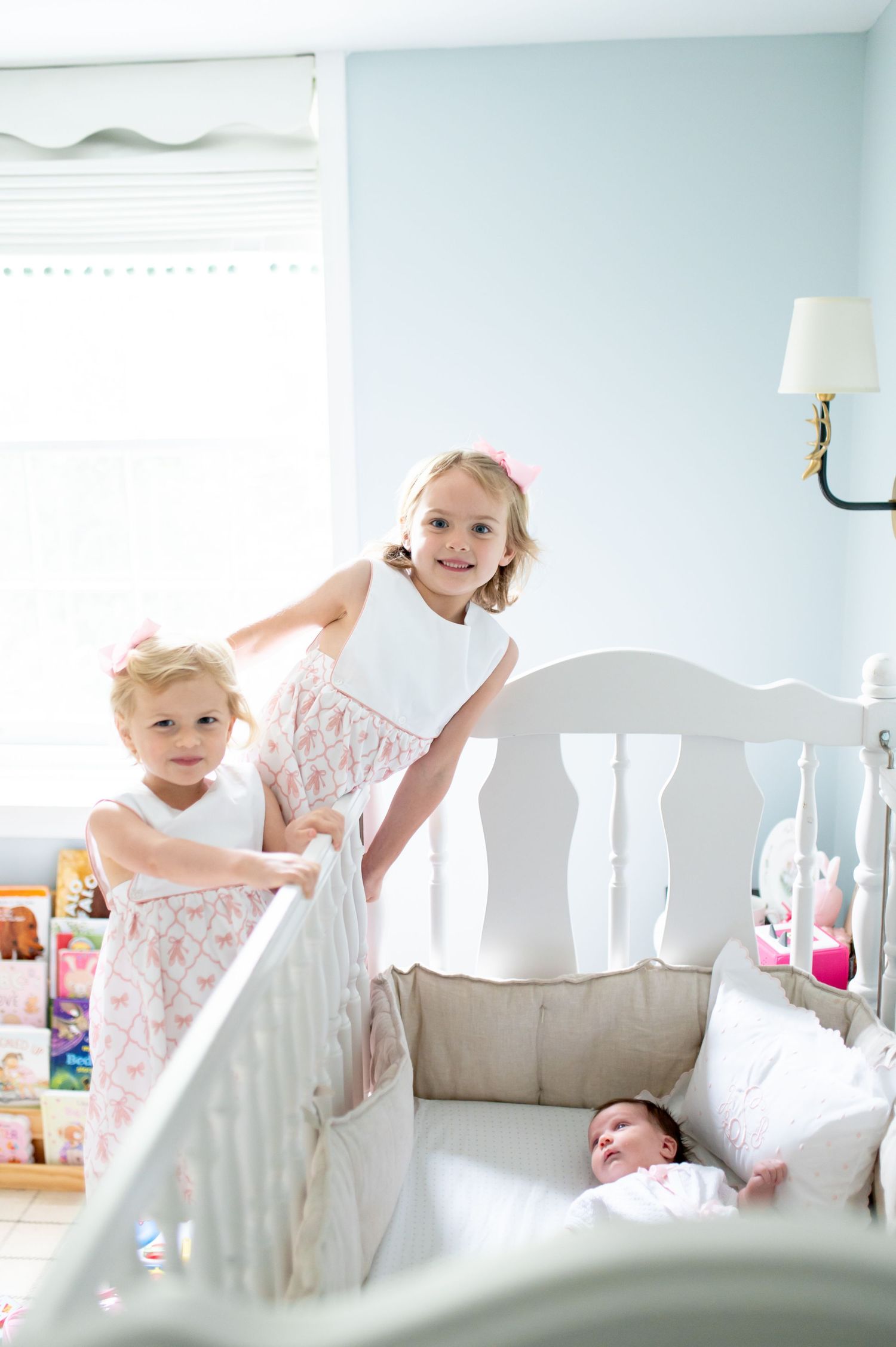 Two young girls in matching pink dresses stand beside a white crib where a newborn baby sleeps in a nursery.