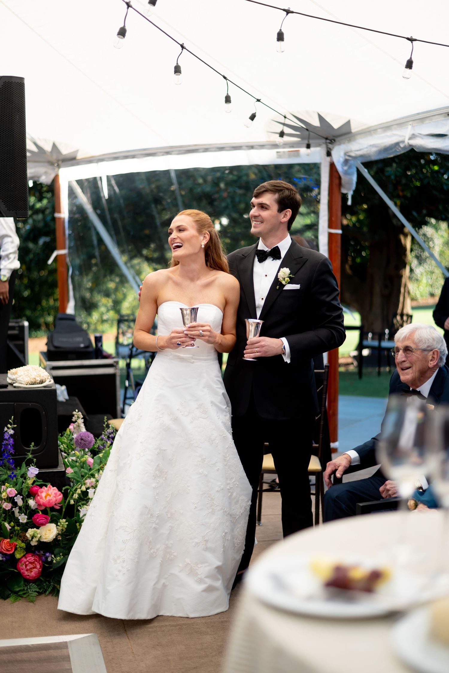 Bride and groom laugh together during wedding reception toast under a tented outdoor venue.