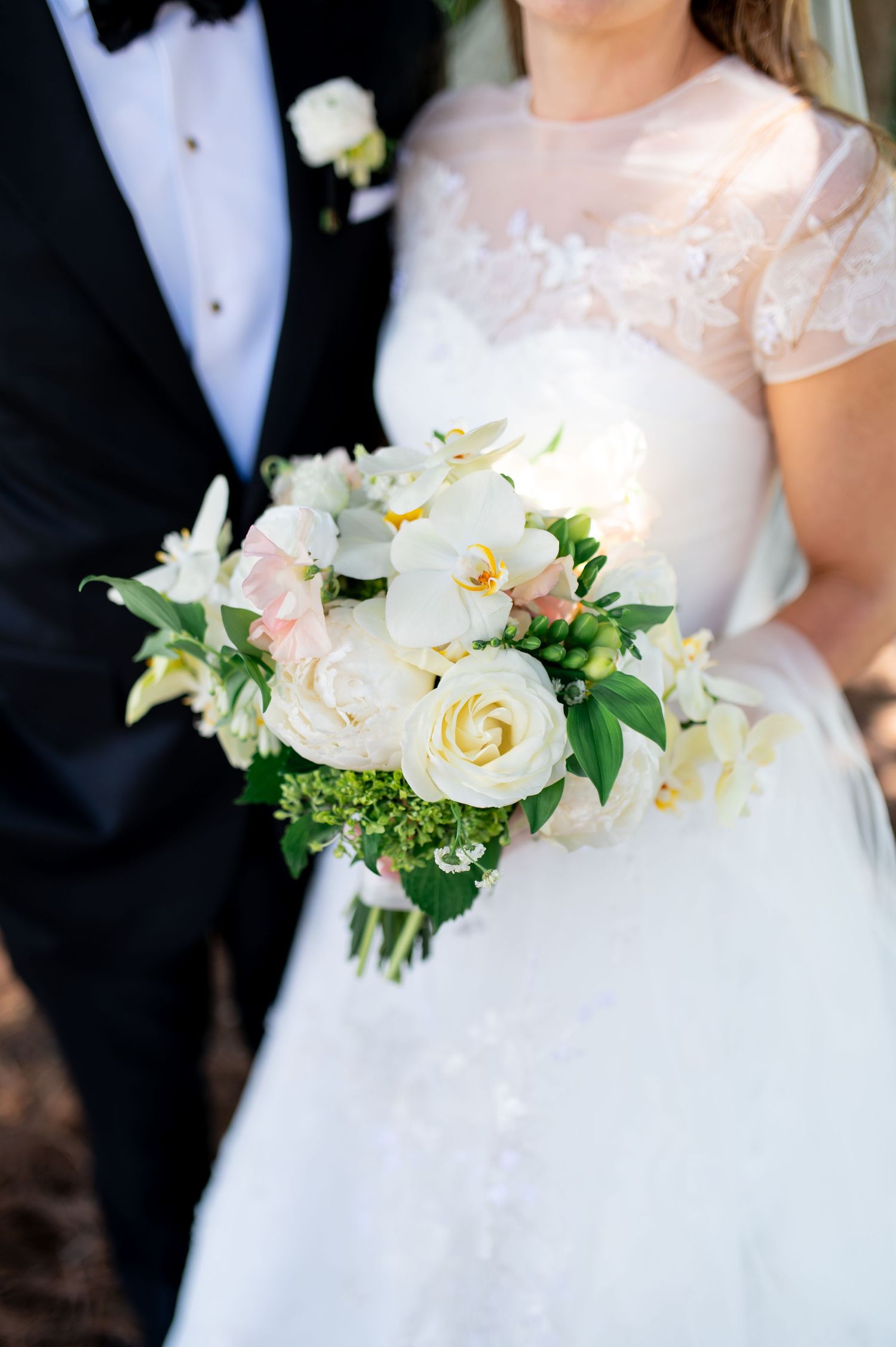 Bride holds elegant white and blush bouquet of roses and orchids while standing beside groom in black tuxedo.