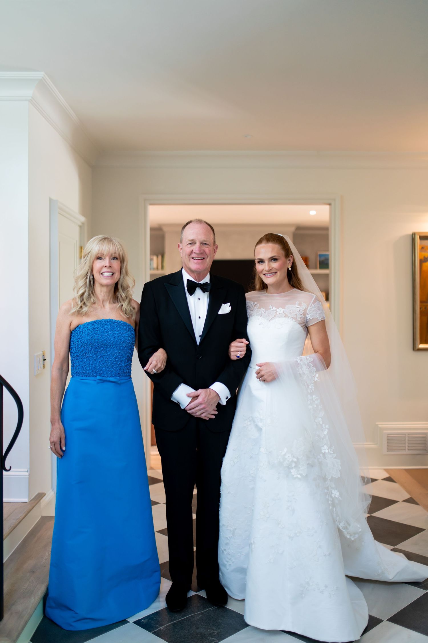 Bride in white lace gown poses with parents in tuxedo and blue gown before wedding ceremony.